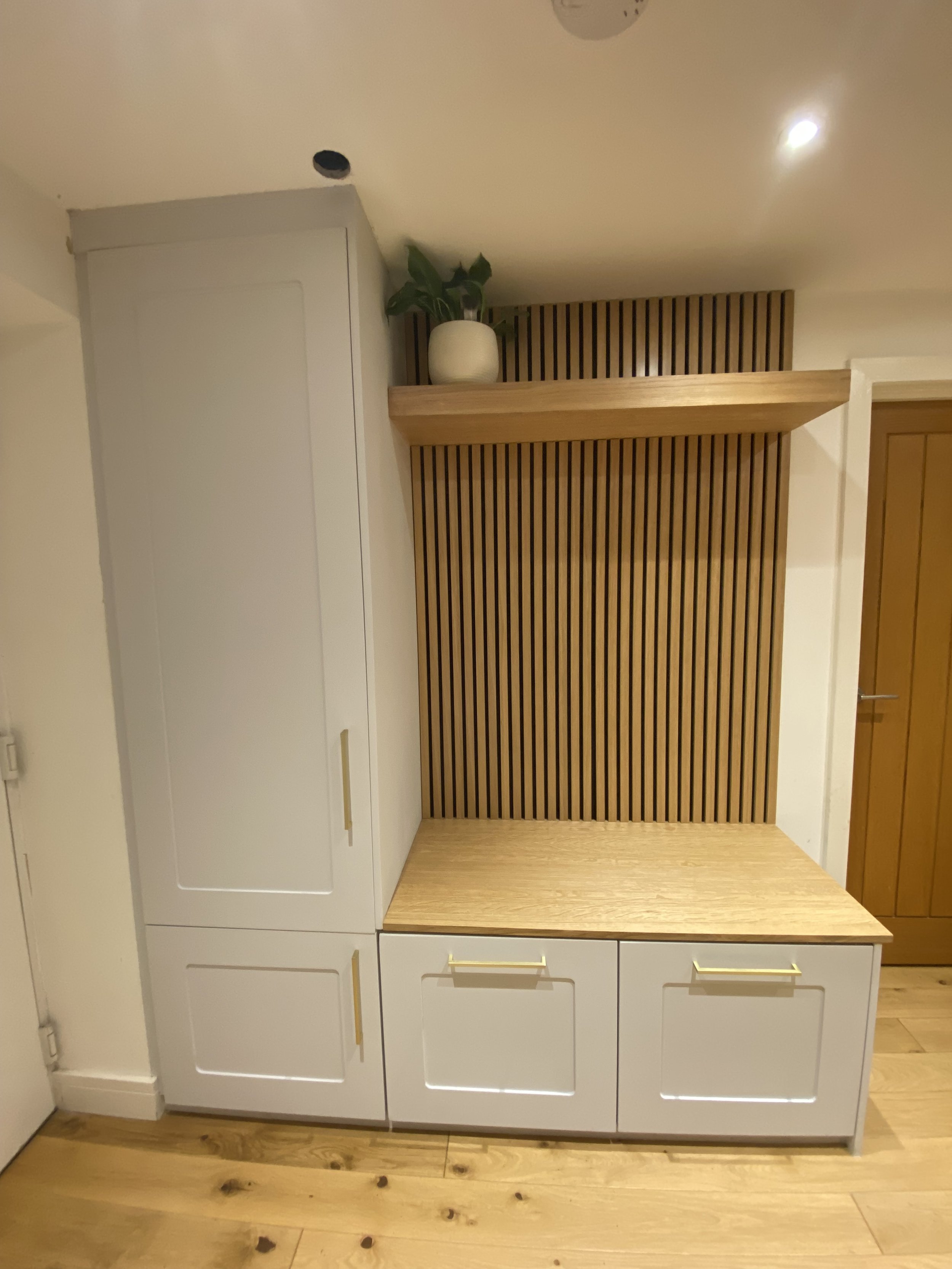 An interior corner with white built-in cabinets, a wooden countertop, a wooden slatted backdrop, a floating wooden shelf with a white pot and green plant, and a wooden door on the right.