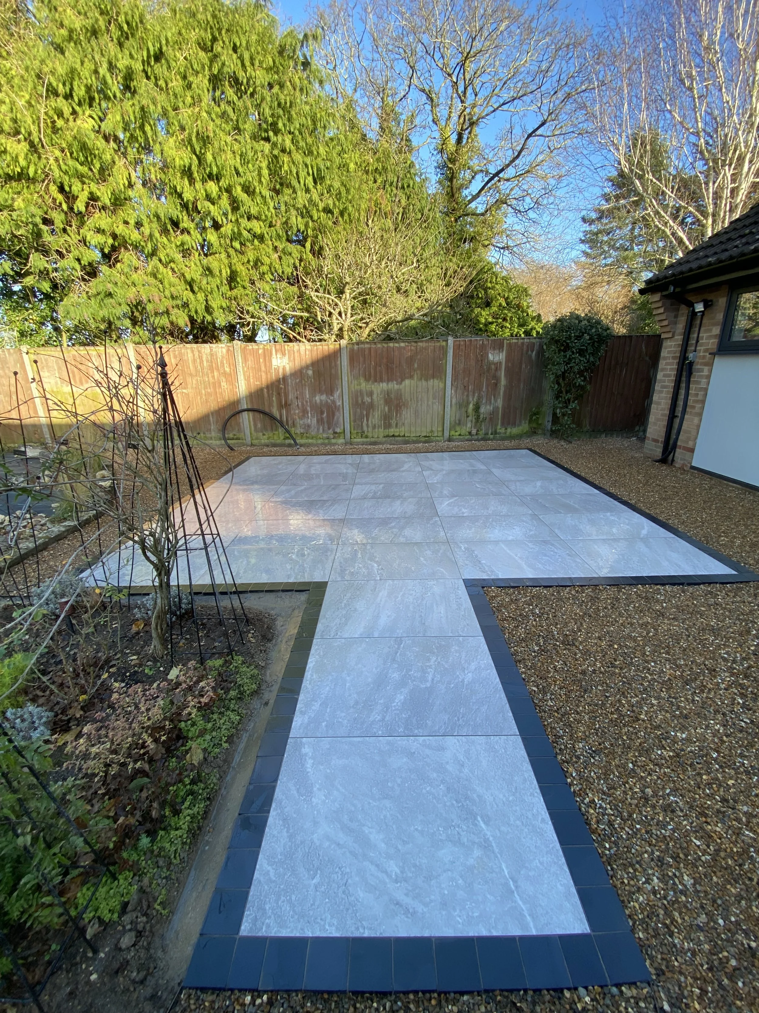Backyard patio with newly installed white tile flooring, bordered by black tiles, next to gravel pathways, with trees and a wooden fence in the background during daytime.