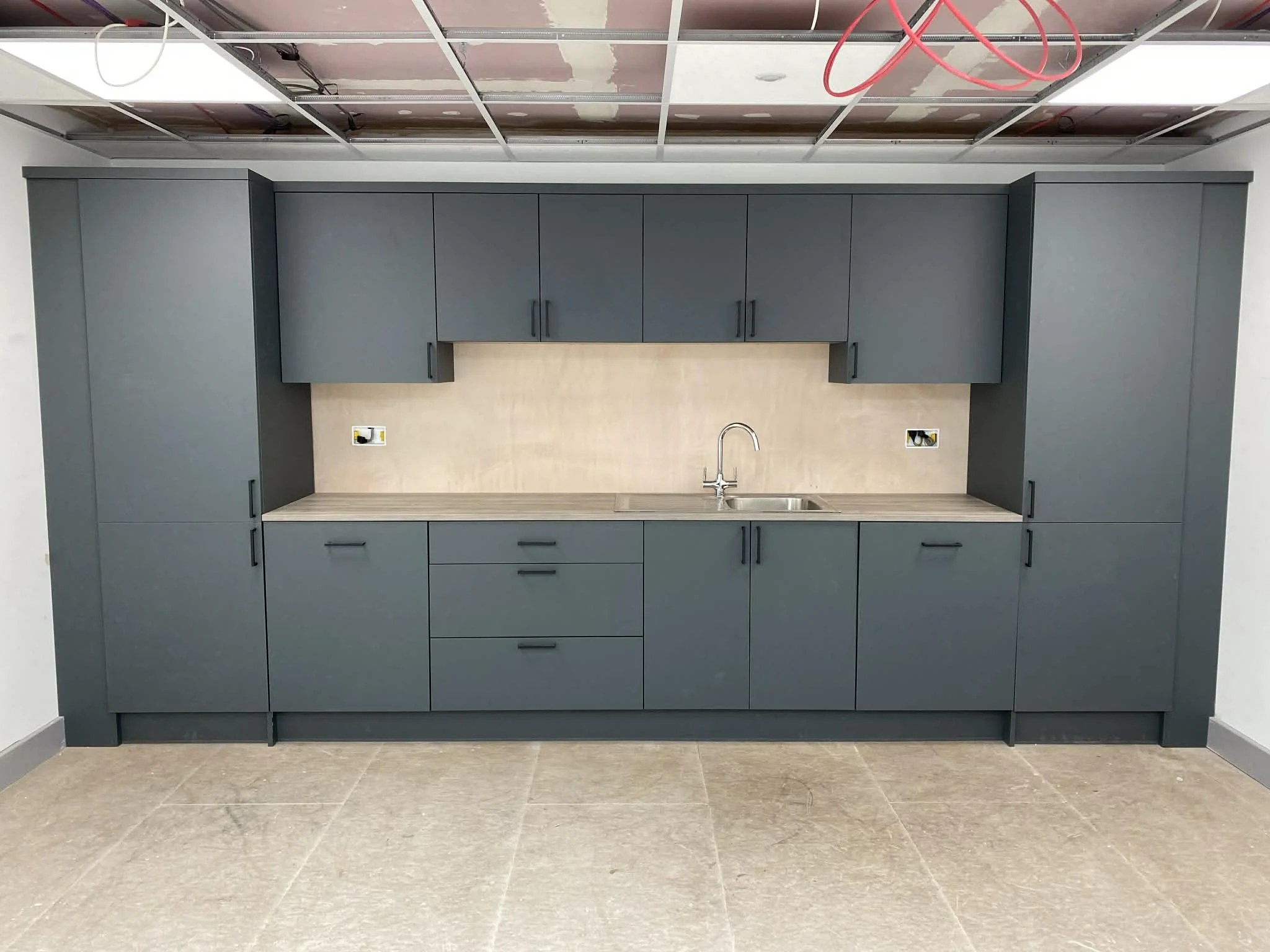 An unfinished kitchen with upper and lower gray cabinets, a beige backsplash, and a single sink with a faucet. Exposed electrical outlets and a construction ceiling with red wiring are visible.