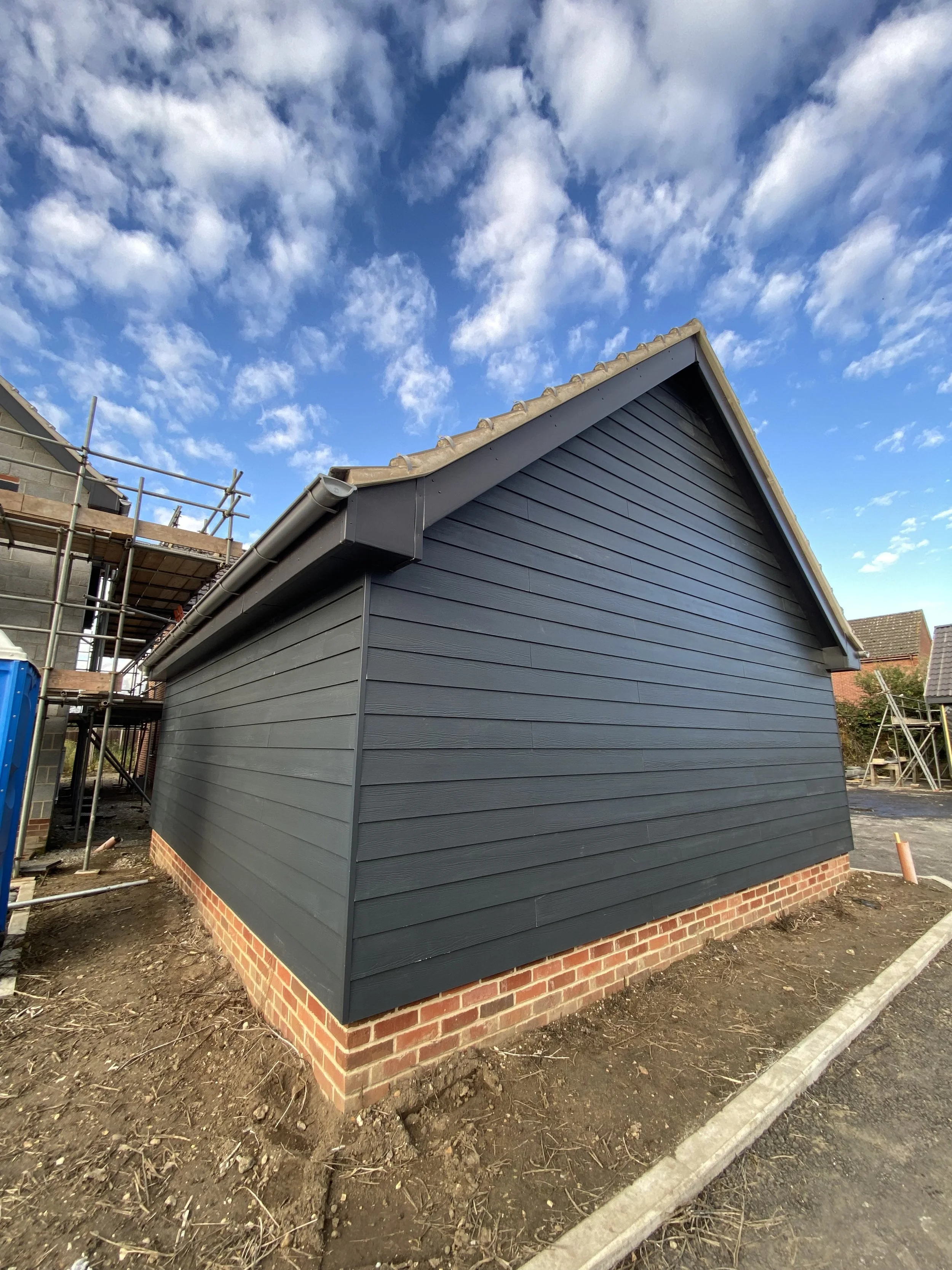 Side view of a newly painted dark gray house with brick foundation, under a partly cloudy blue sky.