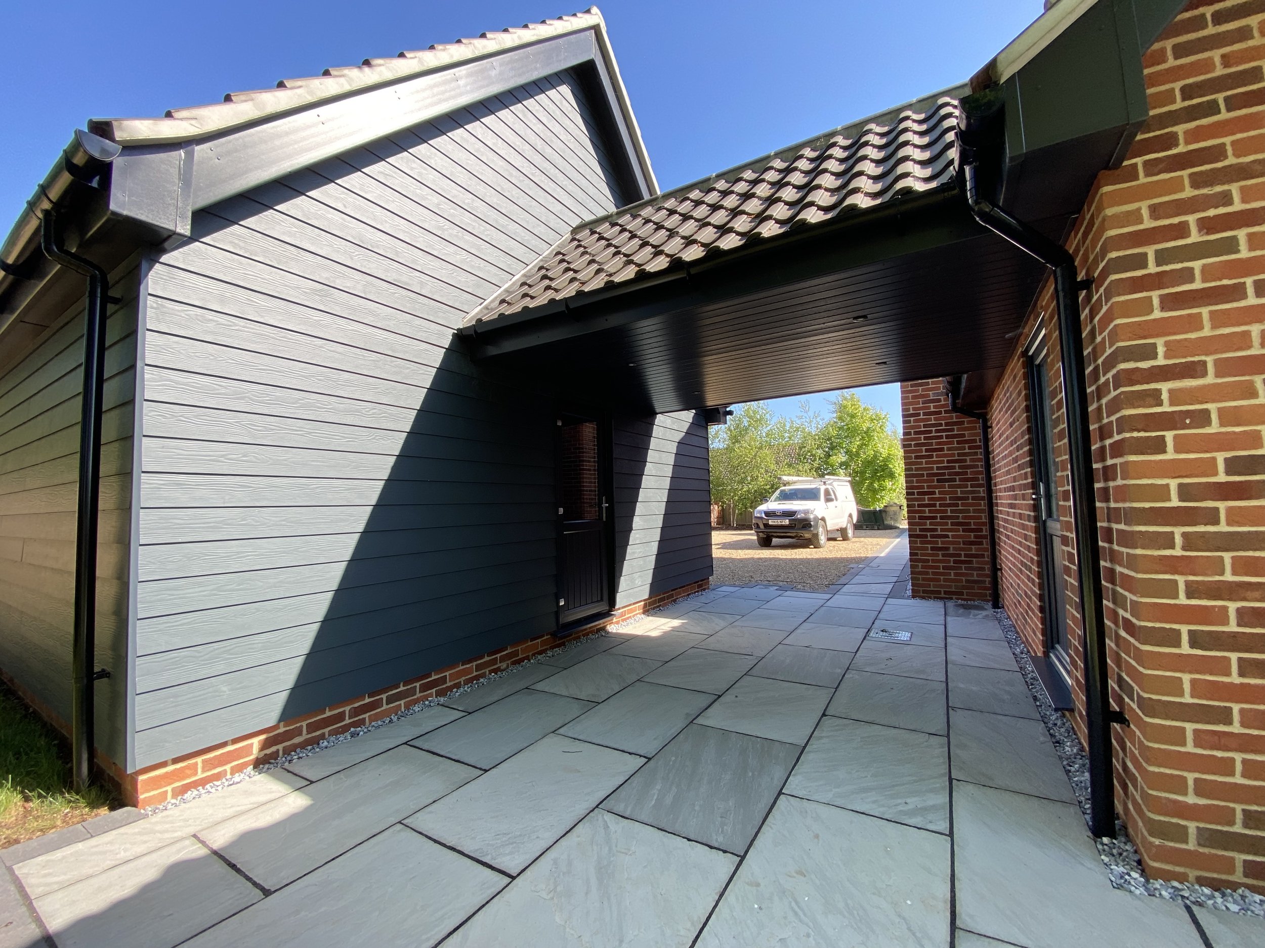 Modern house exterior with a stone walkway, brick wall, and a blue siding wall with a black door. There is a covered area with a black ceiling and a gutter system for drainage.