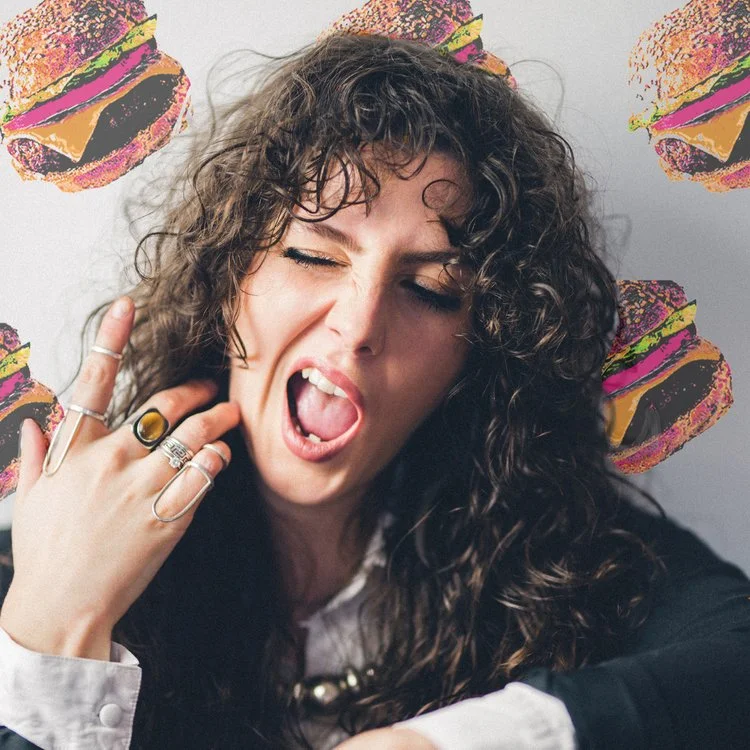 Woman with curly hair posing with her tongue out and eyes closed, wearing rings, gesturing with her hand. Background has colorful illustrations of burgers.