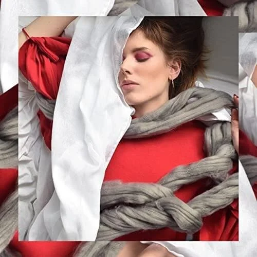 A woman lying with her eyes closed, surrounded by white, gray, and red blankets and sheets, with pink eyeshadow.
