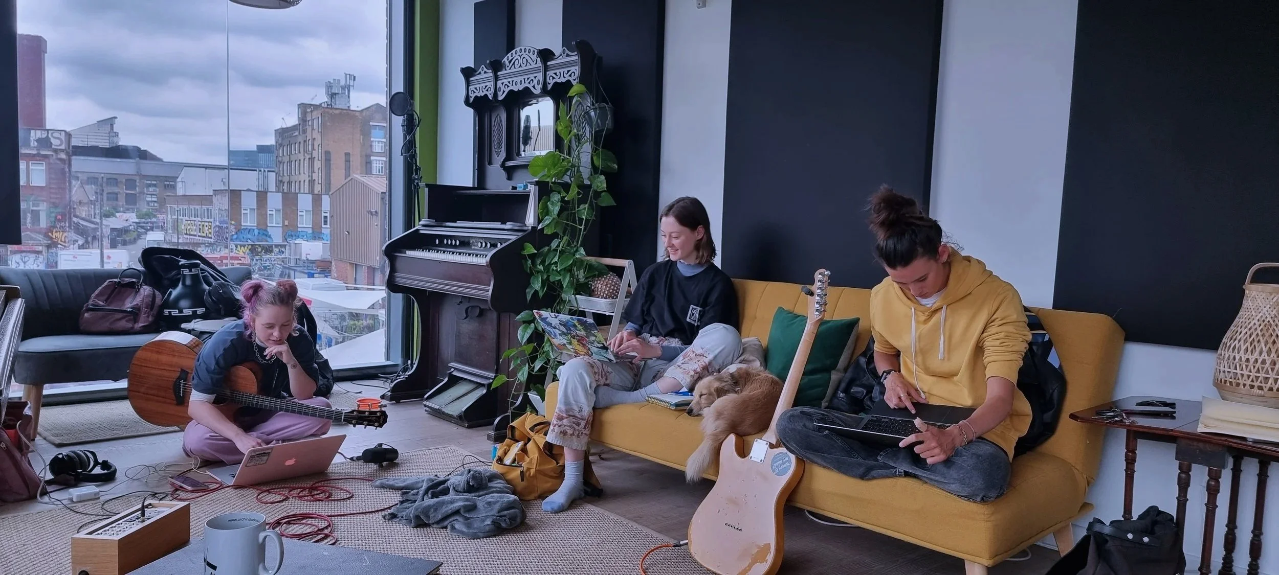 Three young women in a music lounge, two sitting on a yellow couch and one sitting on the floor, engaged with music and devices, with a large window showing city buildings in the background.
