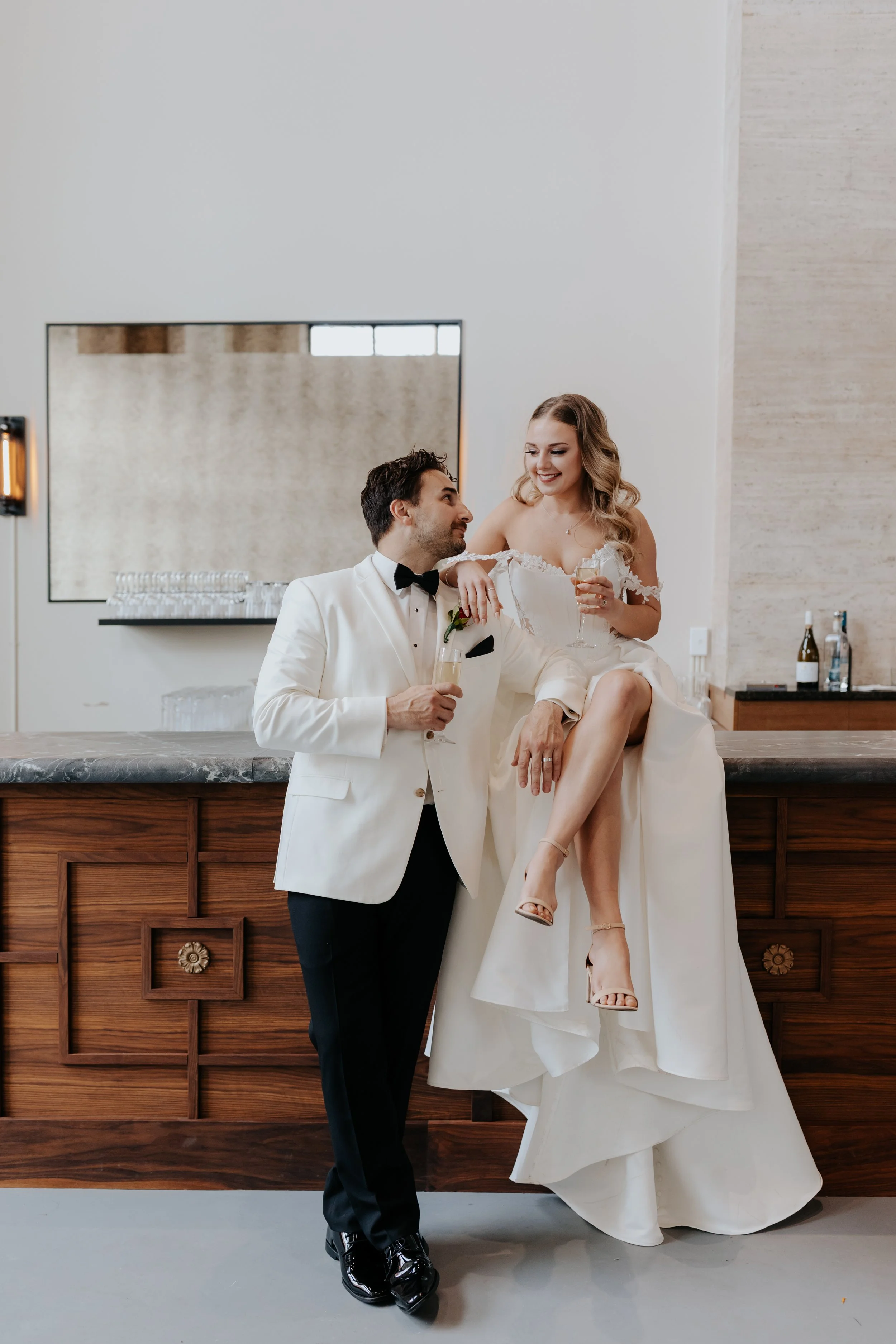 A bride sitting on a bar counter, holding a champagne glass, smiling at the groom who is standing next to her, holding a champagne glass, both dressed in wedding attire.