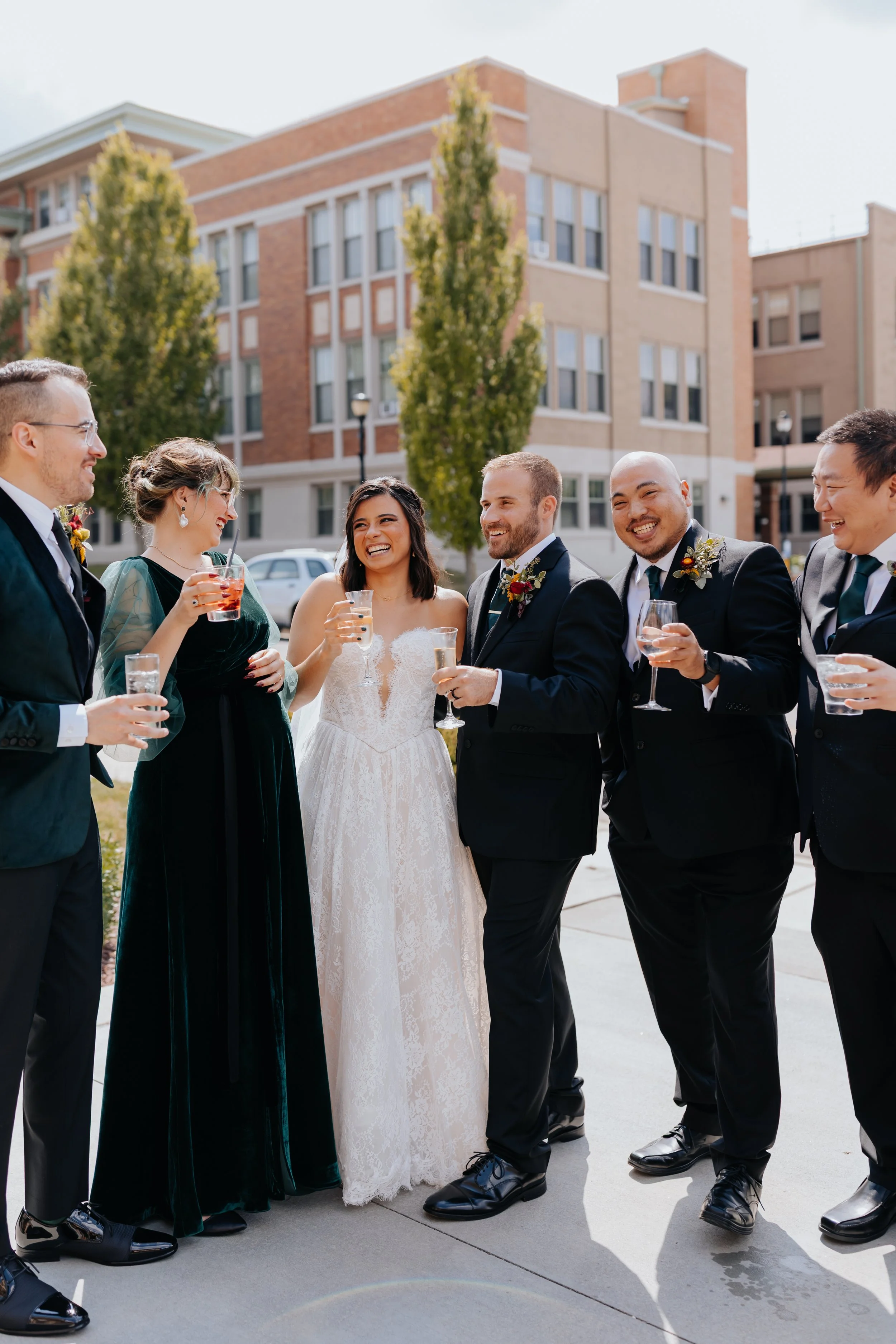 Group of wedding guests dressed in formal attire, laughing and holding drinks outside in front of apartment buildings.