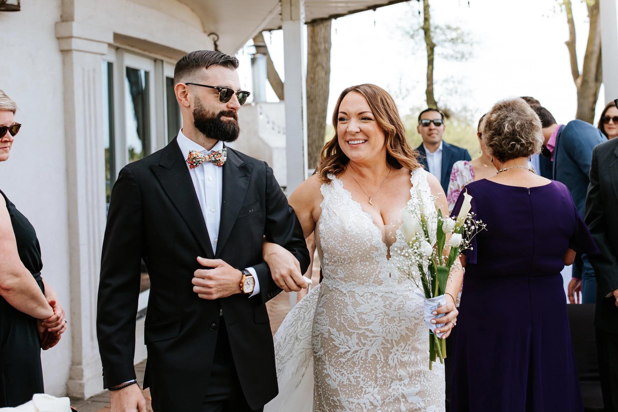 A woman in a white wedding dress smiling and holding a bouquet of white flowers walk arm-in-arm with a man in a black suit with a bow tie and sunglasses. They are outdoors, surrounded by other elegantly dressed guests.