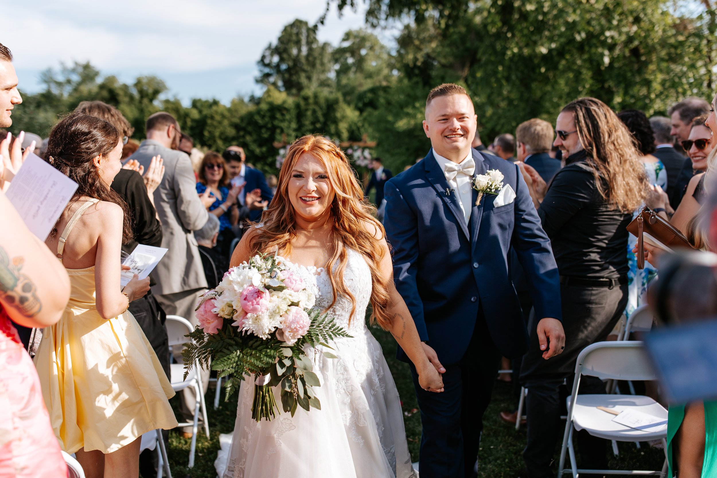 A bride and groom holding hands, smiling as they walk down the aisle at an outdoor wedding, surrounded by guests clapping and celebrating on a sunny day with green trees in the background.