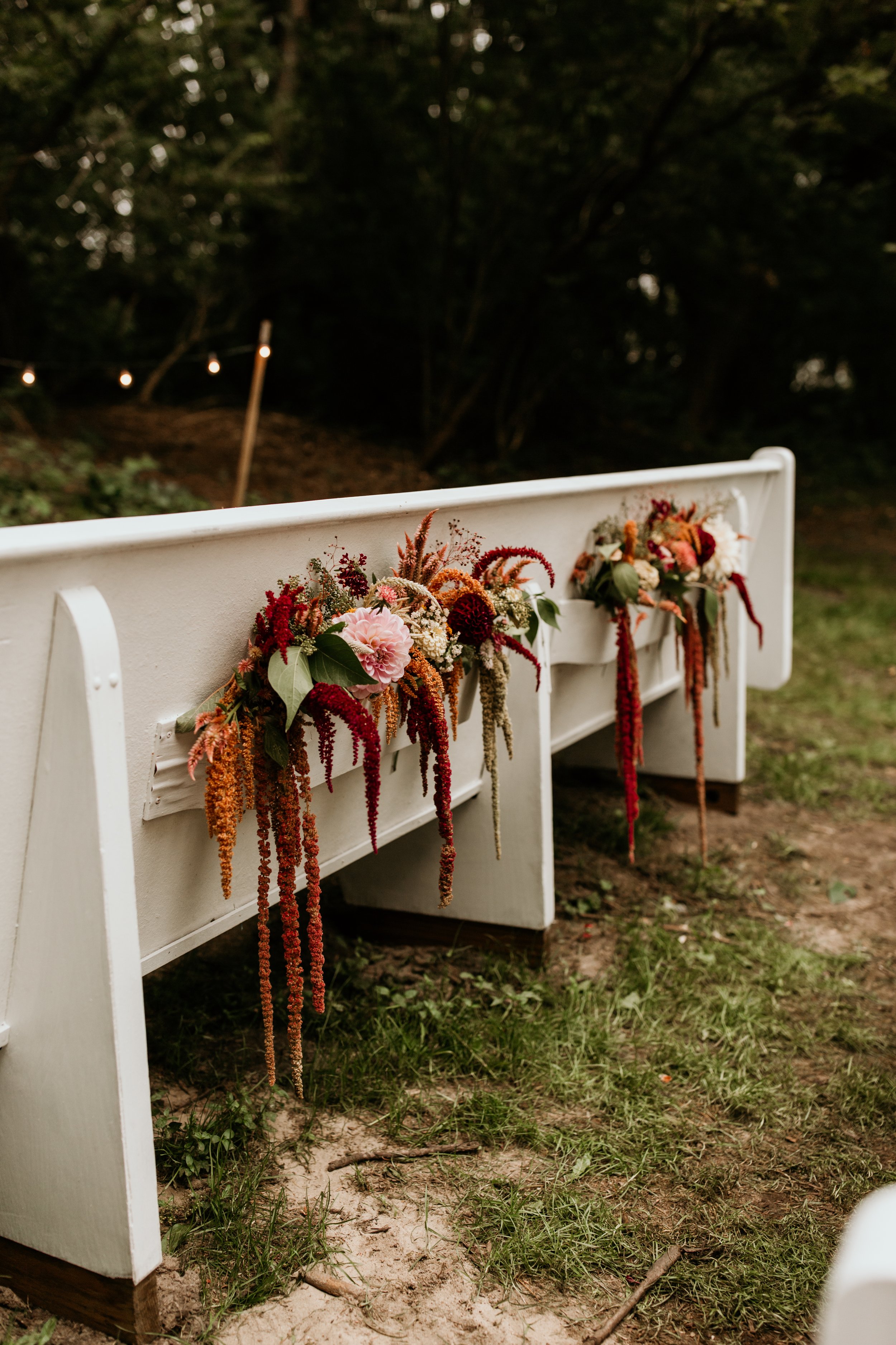Decorative floral arrangements hanging on a white wooden church pew at an outdoor wedding ceremony, with trees and string lights in the background.