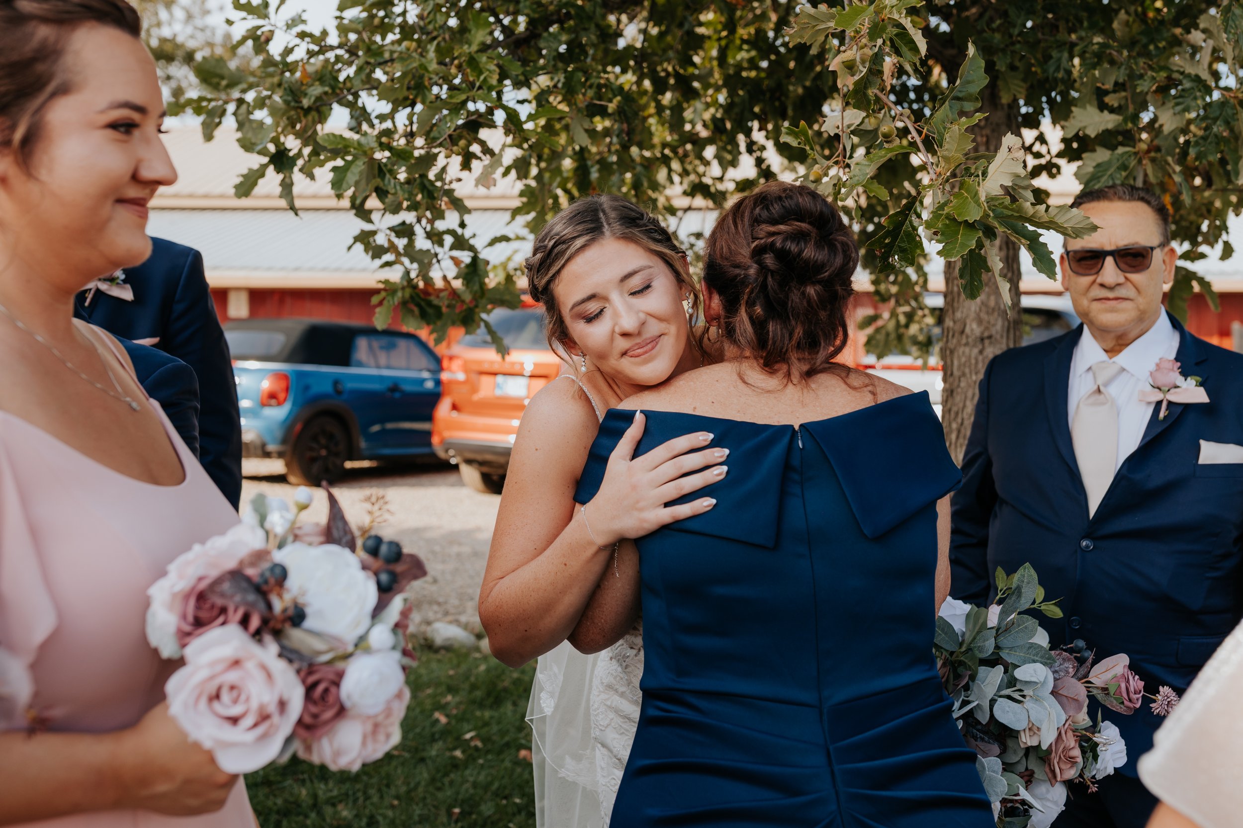 A bride embracing a woman in a navy dress at a wedding outdoor gathering, with bridesmaids holding bouquets and a man in suit with sunglasses observing, under a tree with cars in the background.