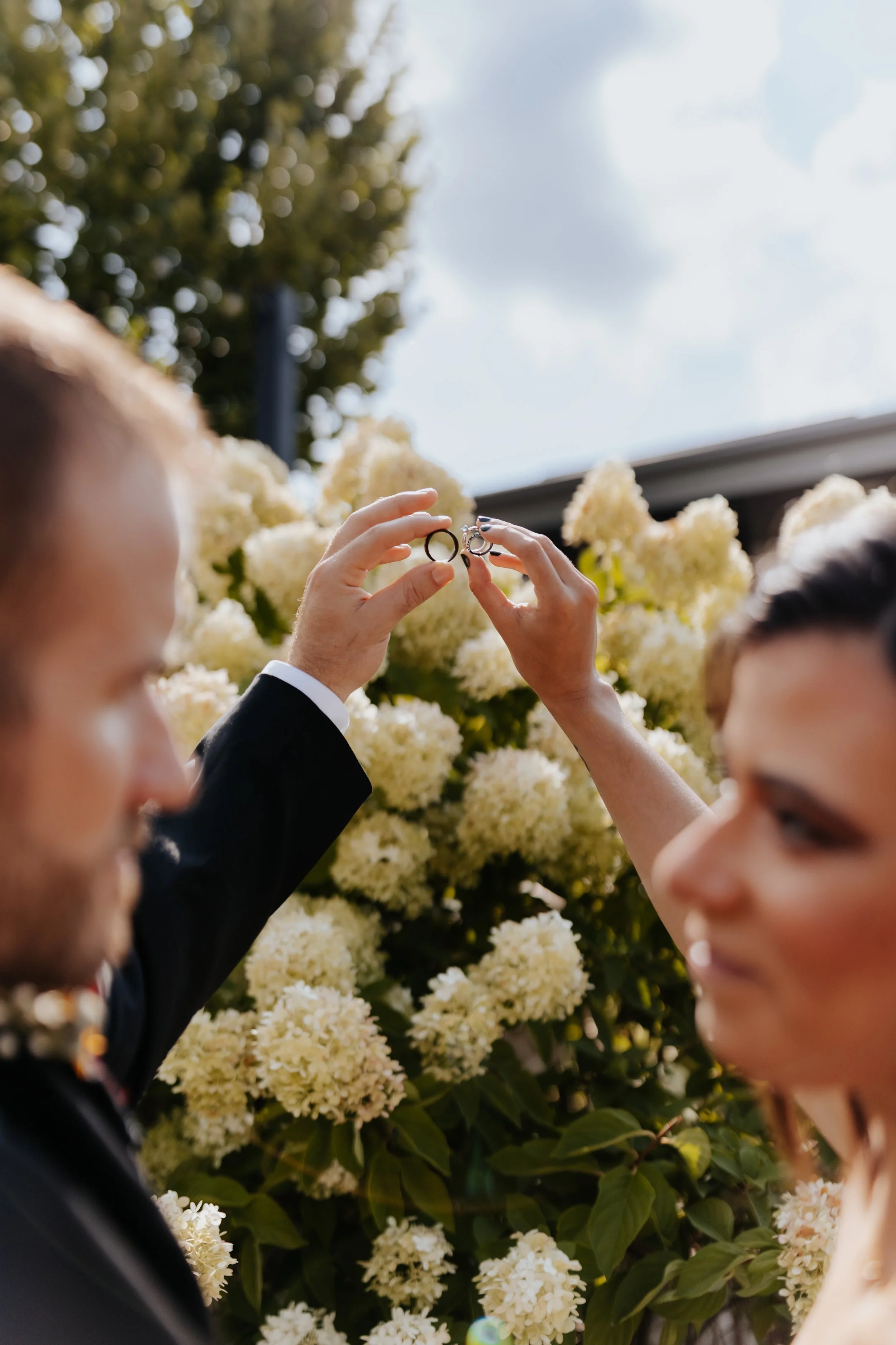 A couple exchanging wedding rings outdoors with white flowers and a cloudy sky in the background.