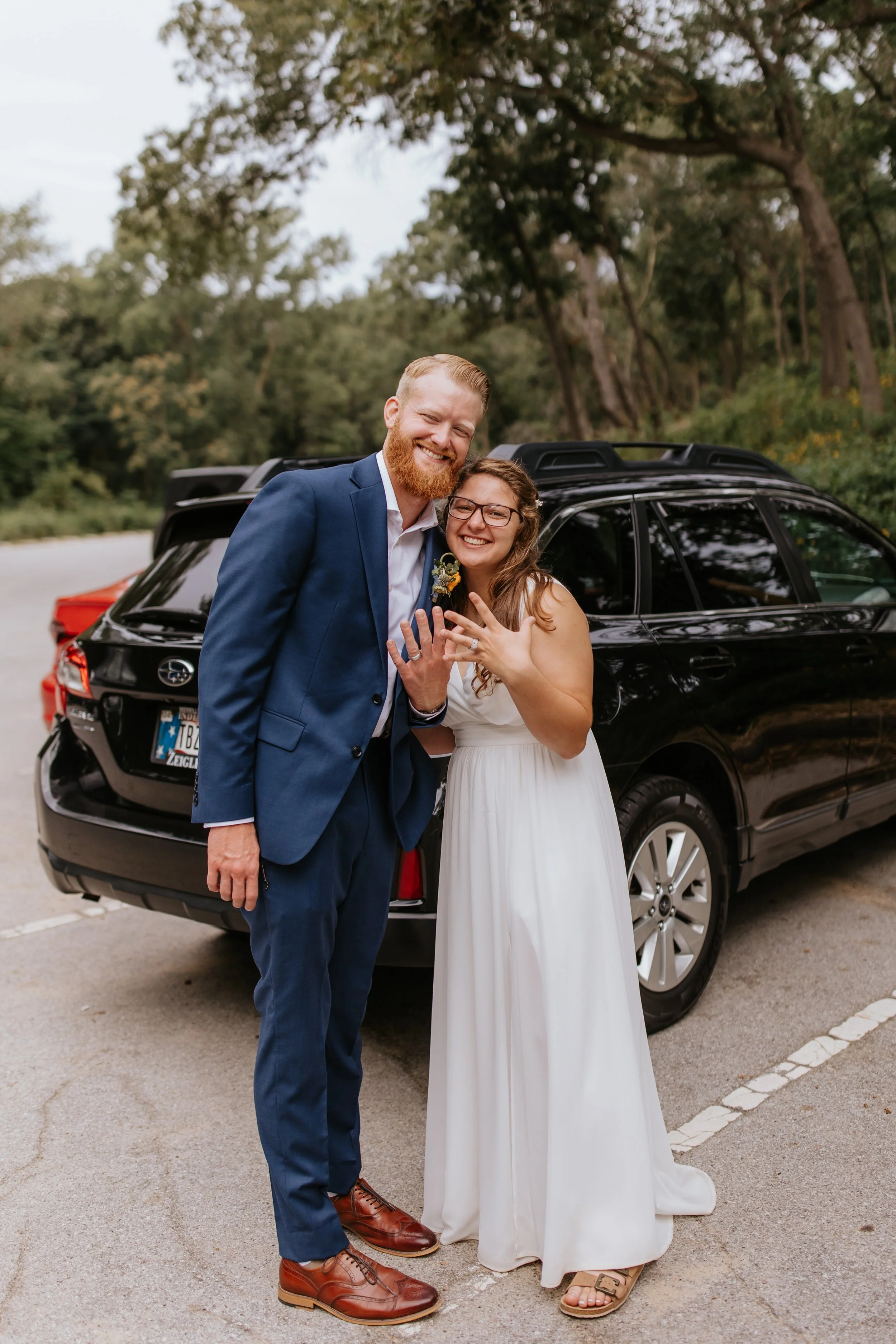 A smiling man in a blue suit and a smiling woman in a white dress show off their rings in front of a black car, with trees and greenery in the background.
