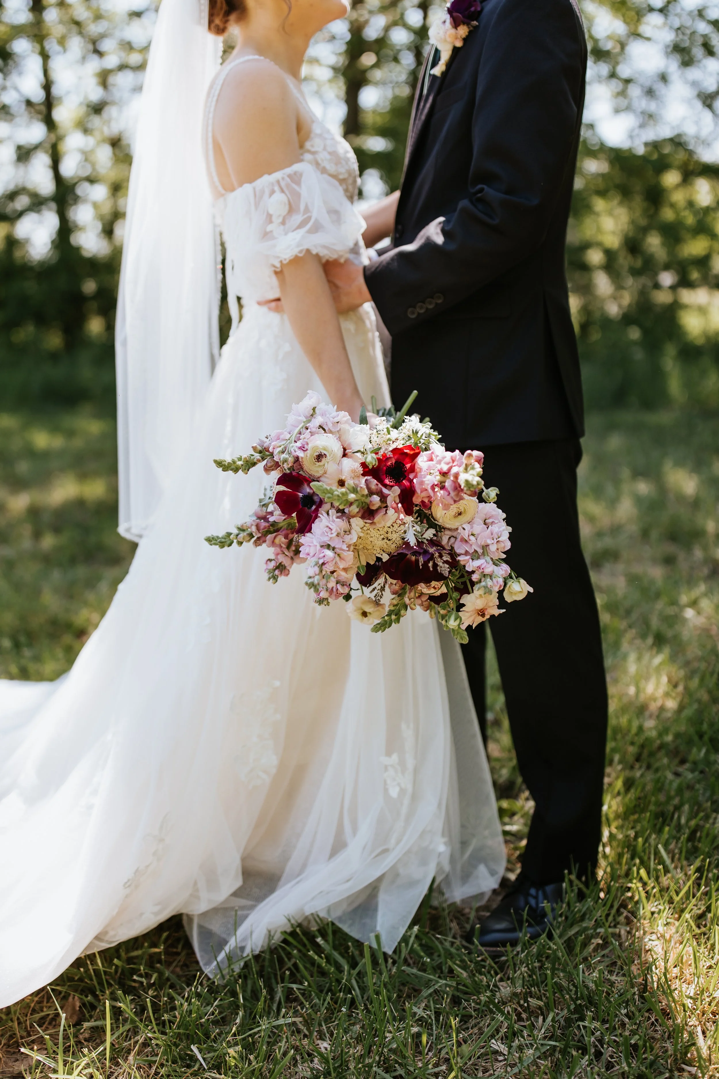 A bride and groom facing each other outdoors, holding hands, with the bride holding a colorful bouquet of flowers; the bride wears a white wedding dress with off-the-shoulder sleeves, and the groom is dressed in a black suit.