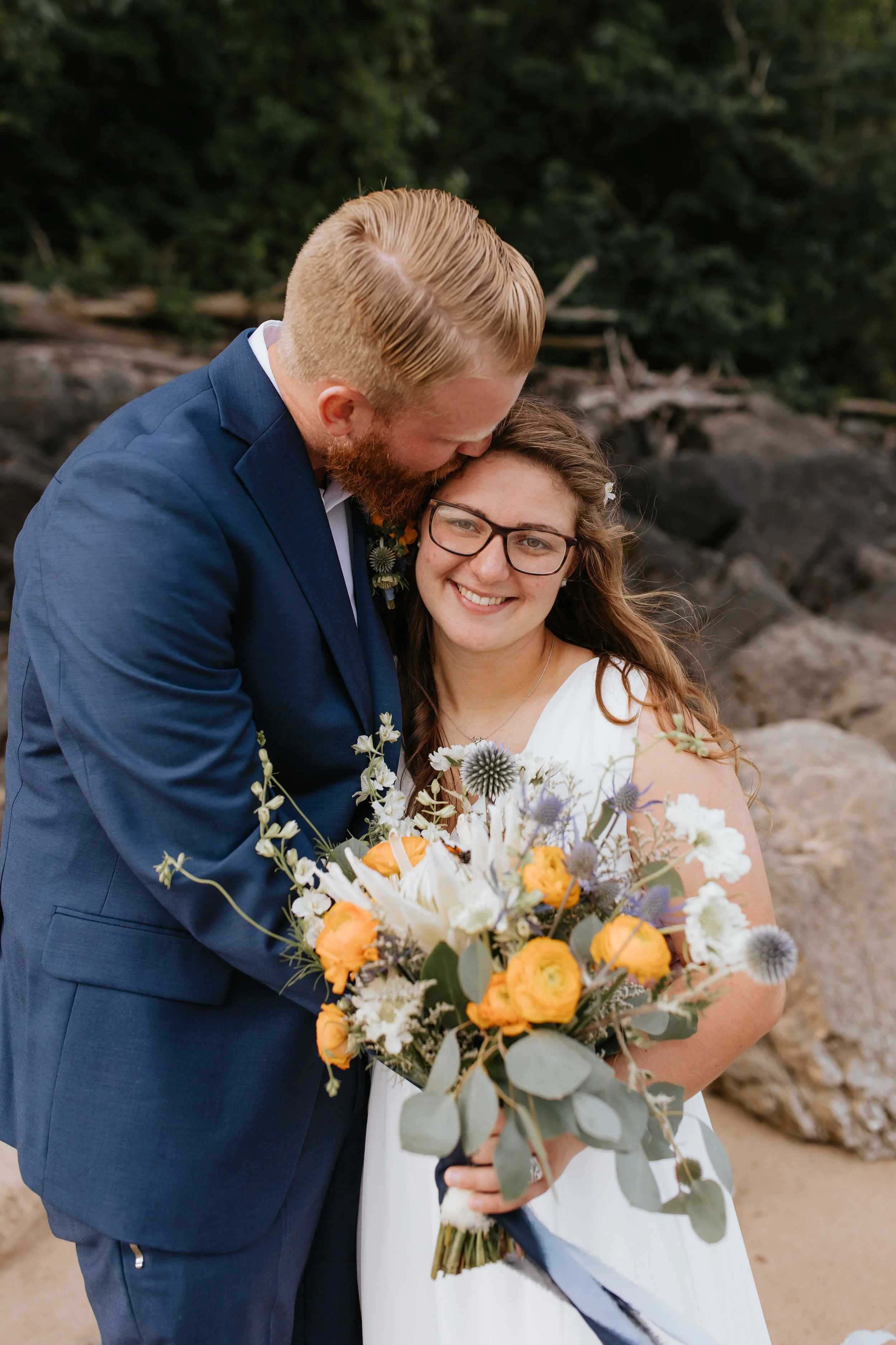 A smiling woman in a white dress holding a bouquet of flowers with a man in a blue suit kissing her on the head.