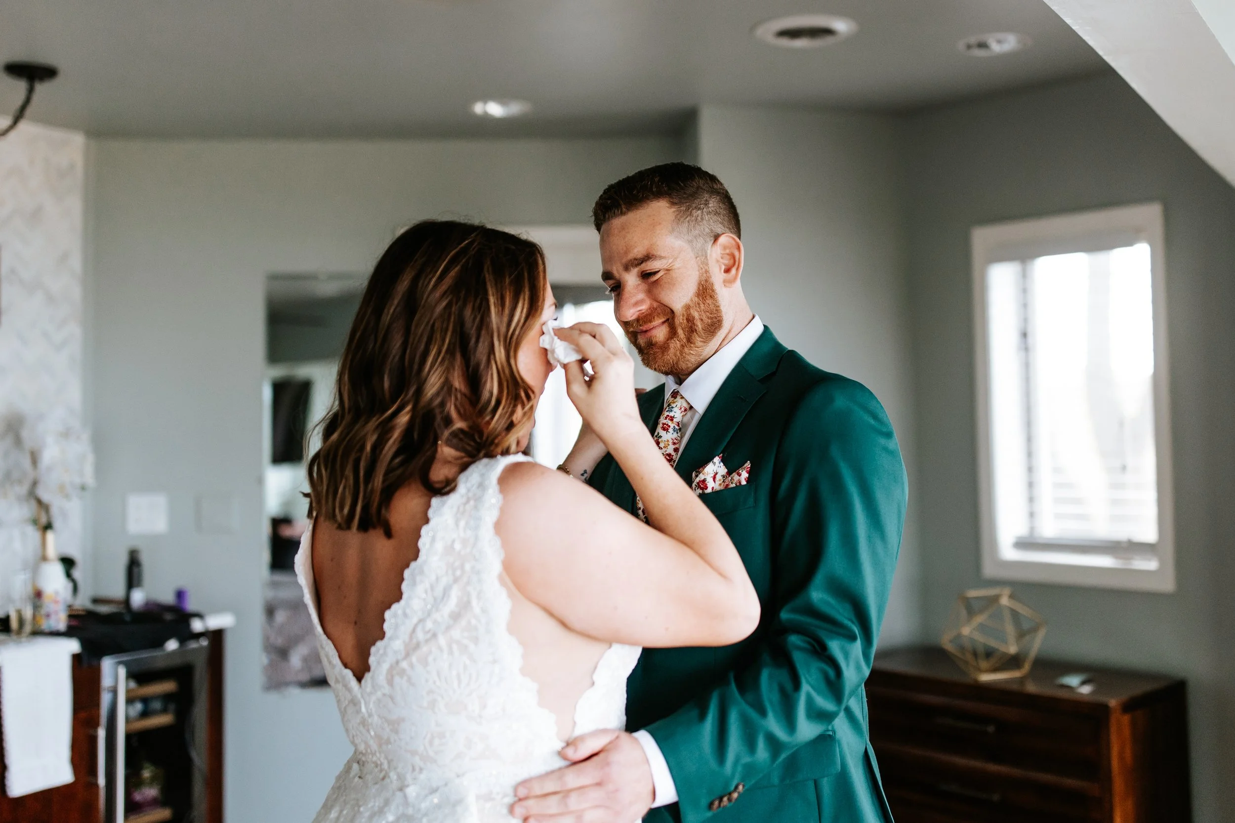 A bride and groom sharing an emotional moment indoors, with the bride wiping tears from her eyes as the groom tenderly holds her. The bride is wearing a white lace wedding dress, and the groom is in a dark green suit with a floral tie and pocket squa