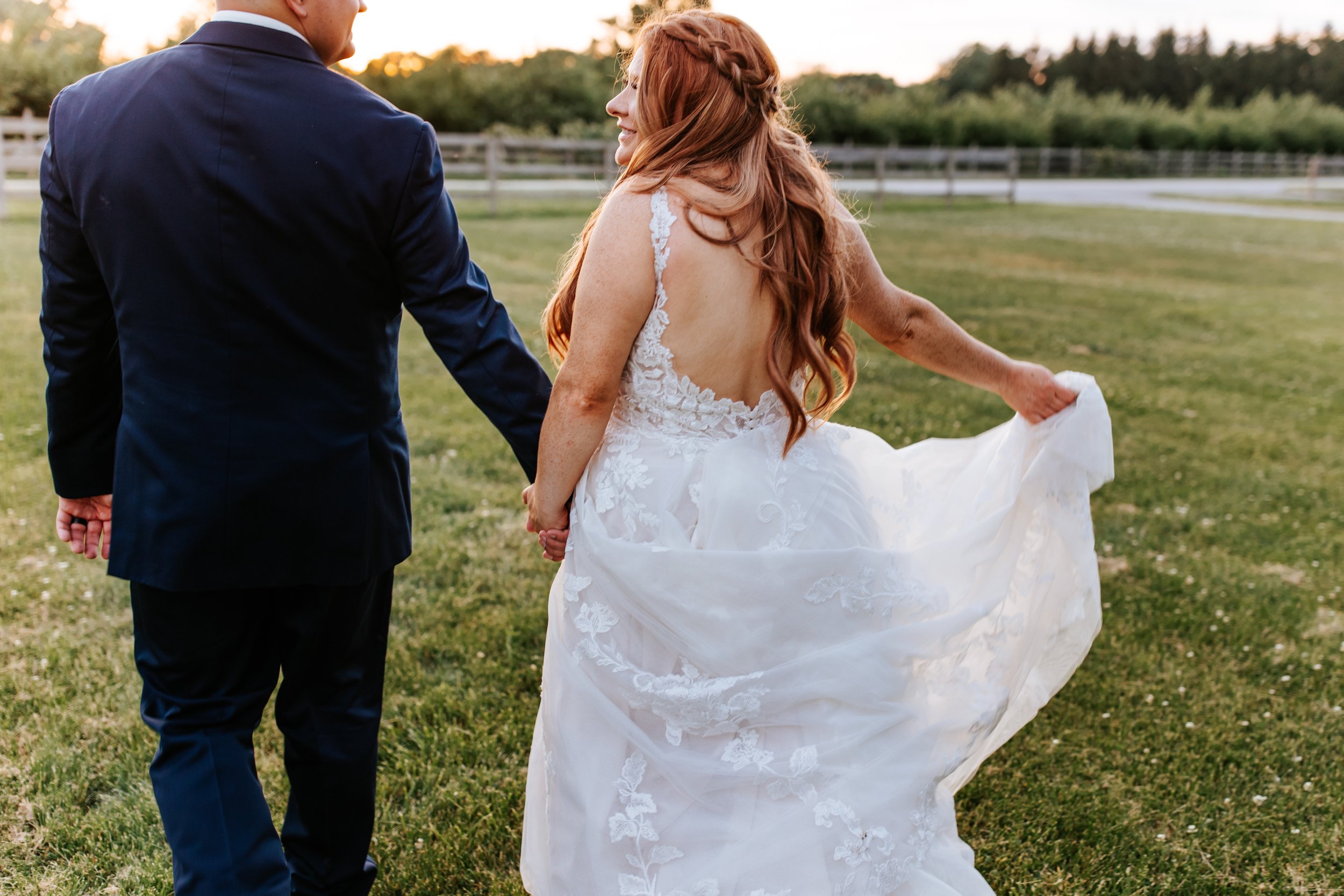 A bride and groom holding hands in a grassy outdoor area at sunset, with the bride lifting her wedding dress slightly as they walk.