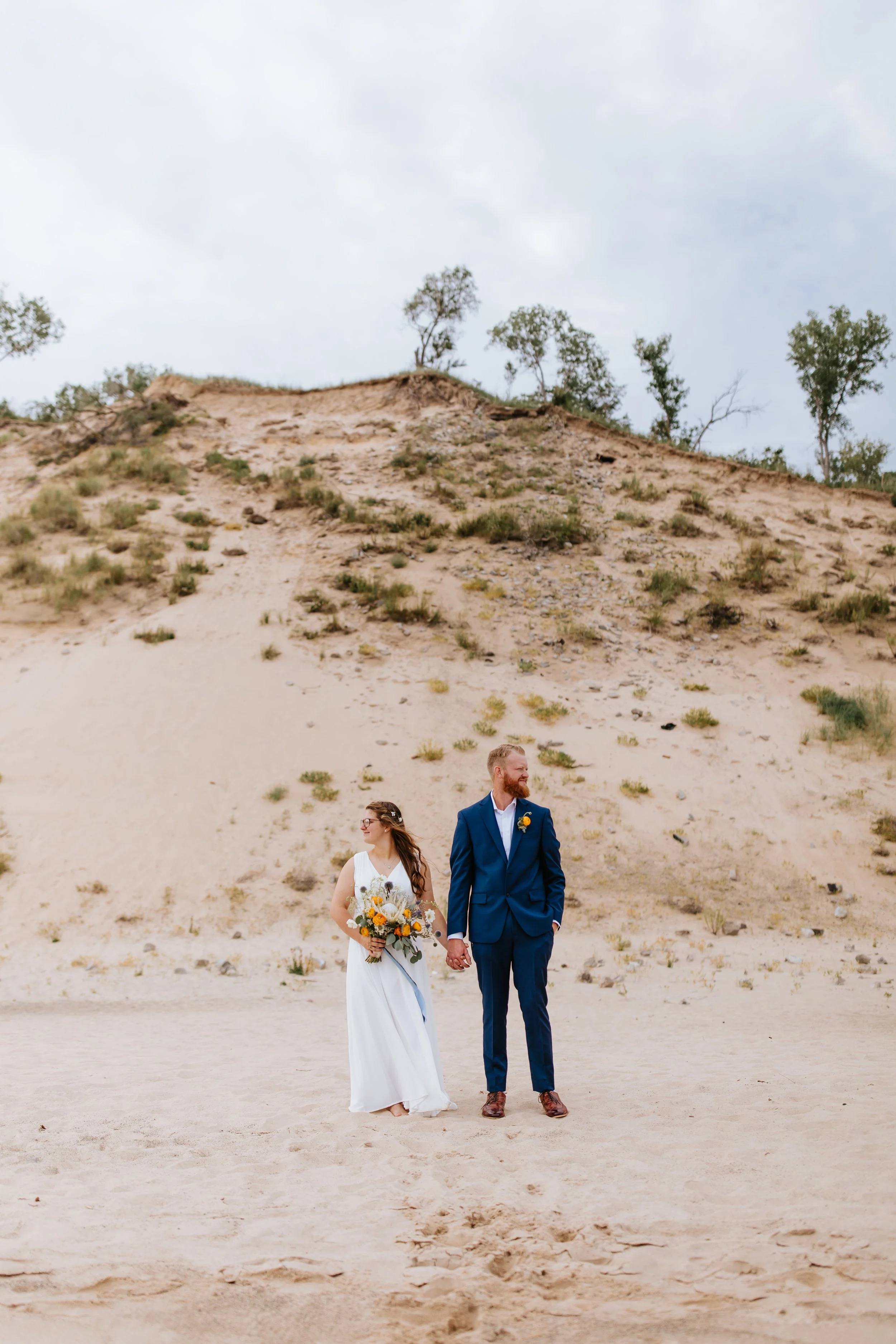 A bride in a white wedding dress holding a bouquet of flowers and a groom in a blue suit holding hands, standing on sandy terrain in front of a hillside with sparse trees under a cloudy sky.