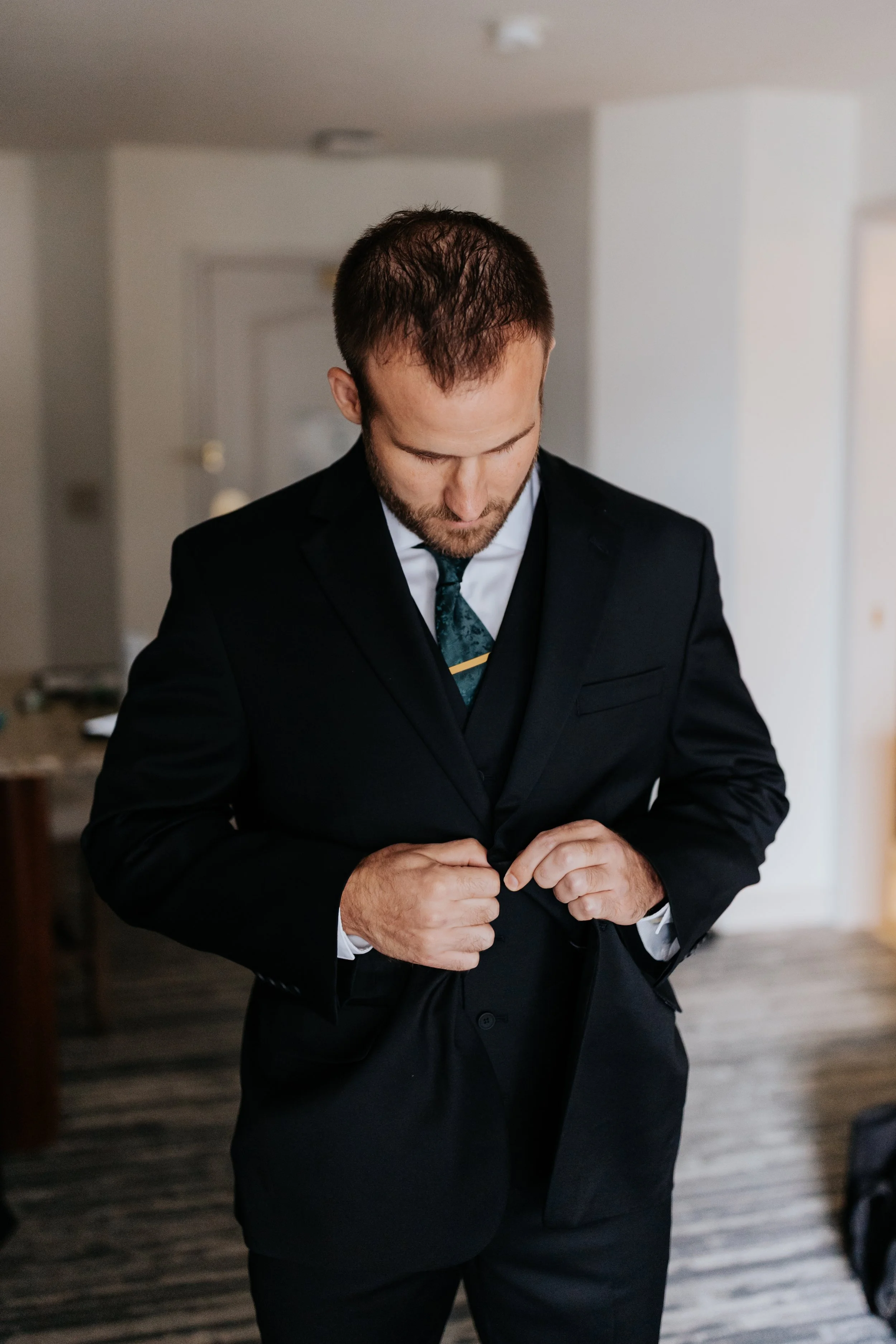 A man in a black suit adjusting his jacket in a hotel room or similar indoor setting.