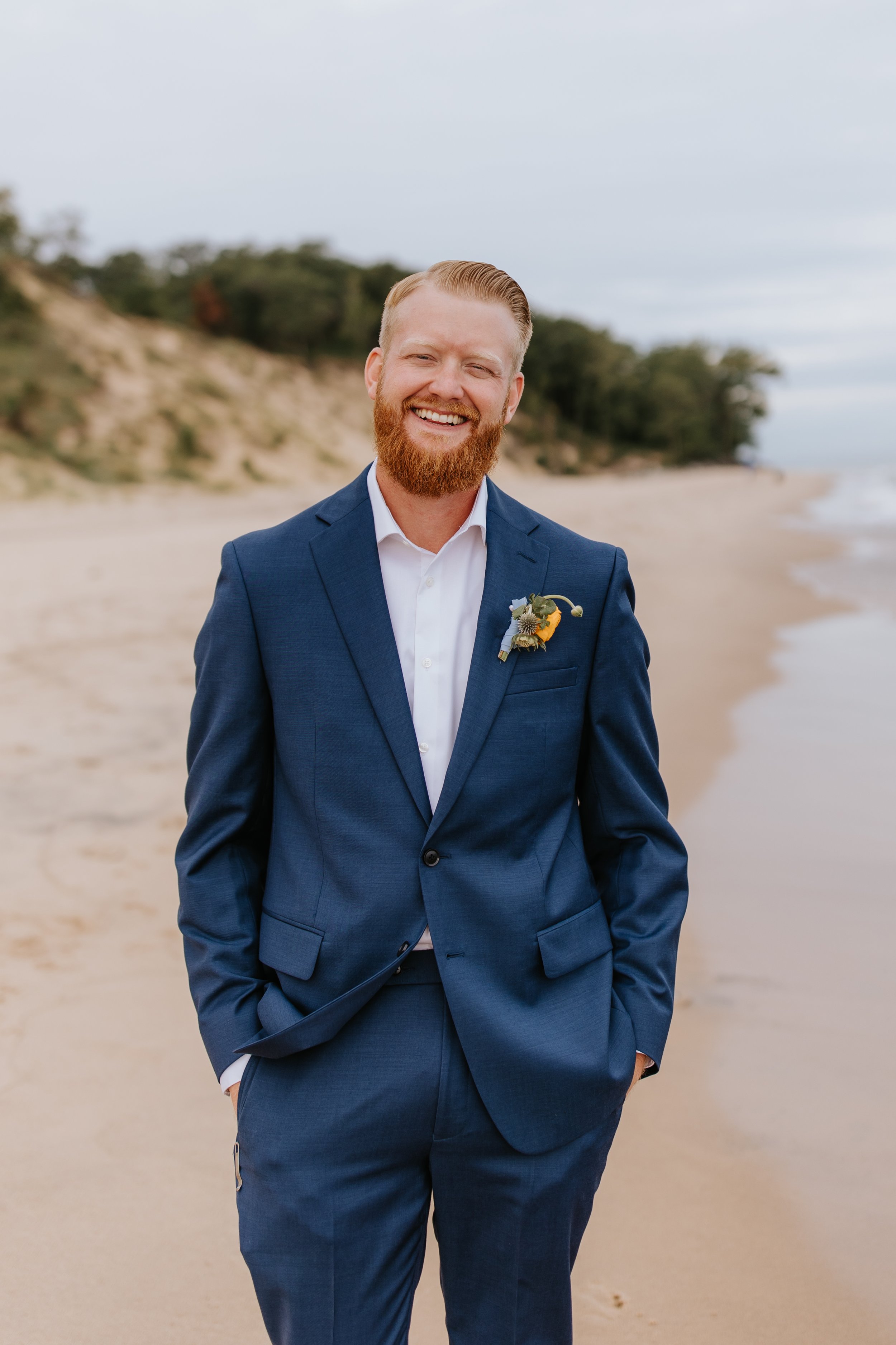 A man in a blue suit standing shirtless on a sandy beach with dunes and trees in the background, smiling at the camera.