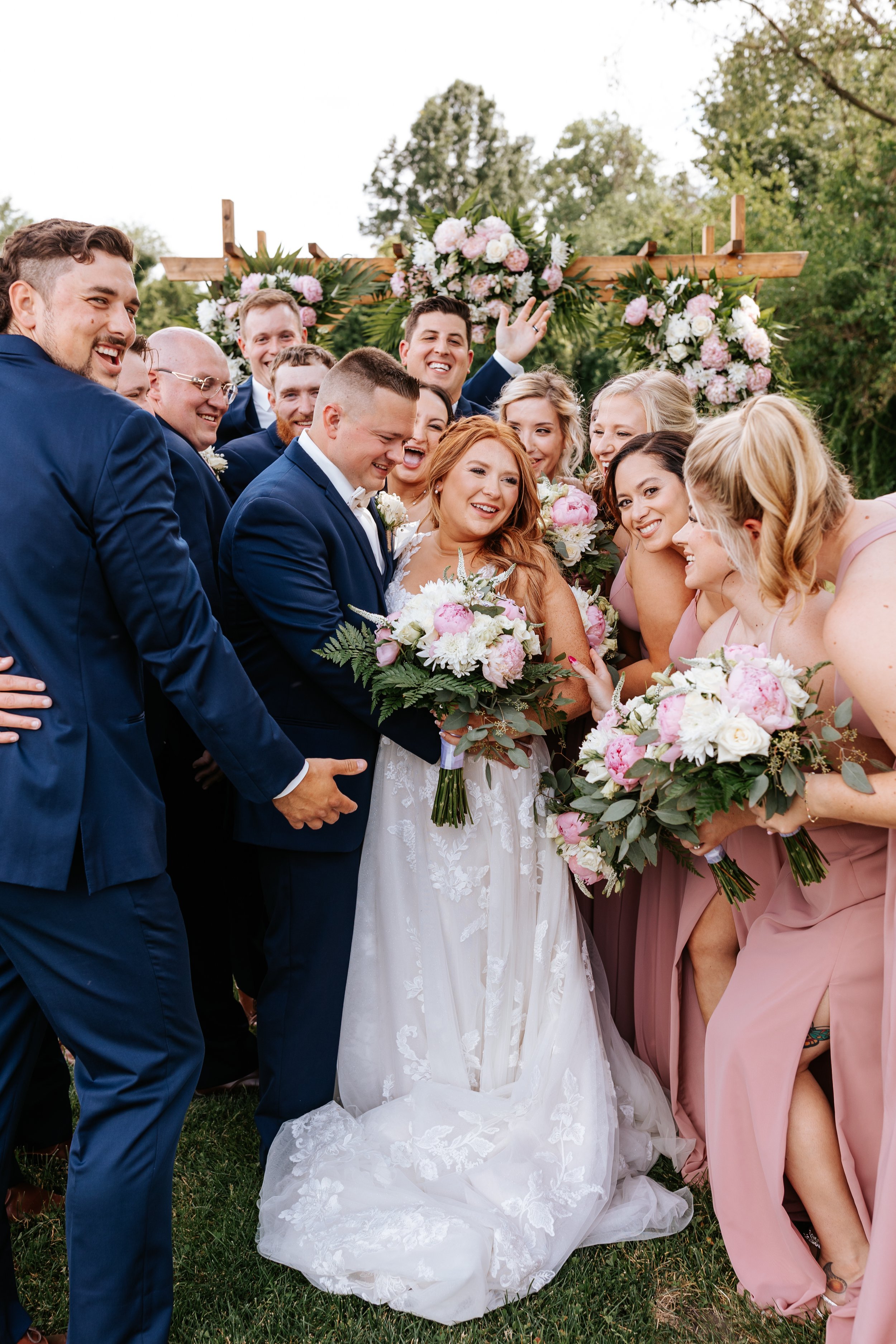 Group of wedding guests gathered around the bride and groom, smiling and holding bouquets, with floral arrangements in the background.