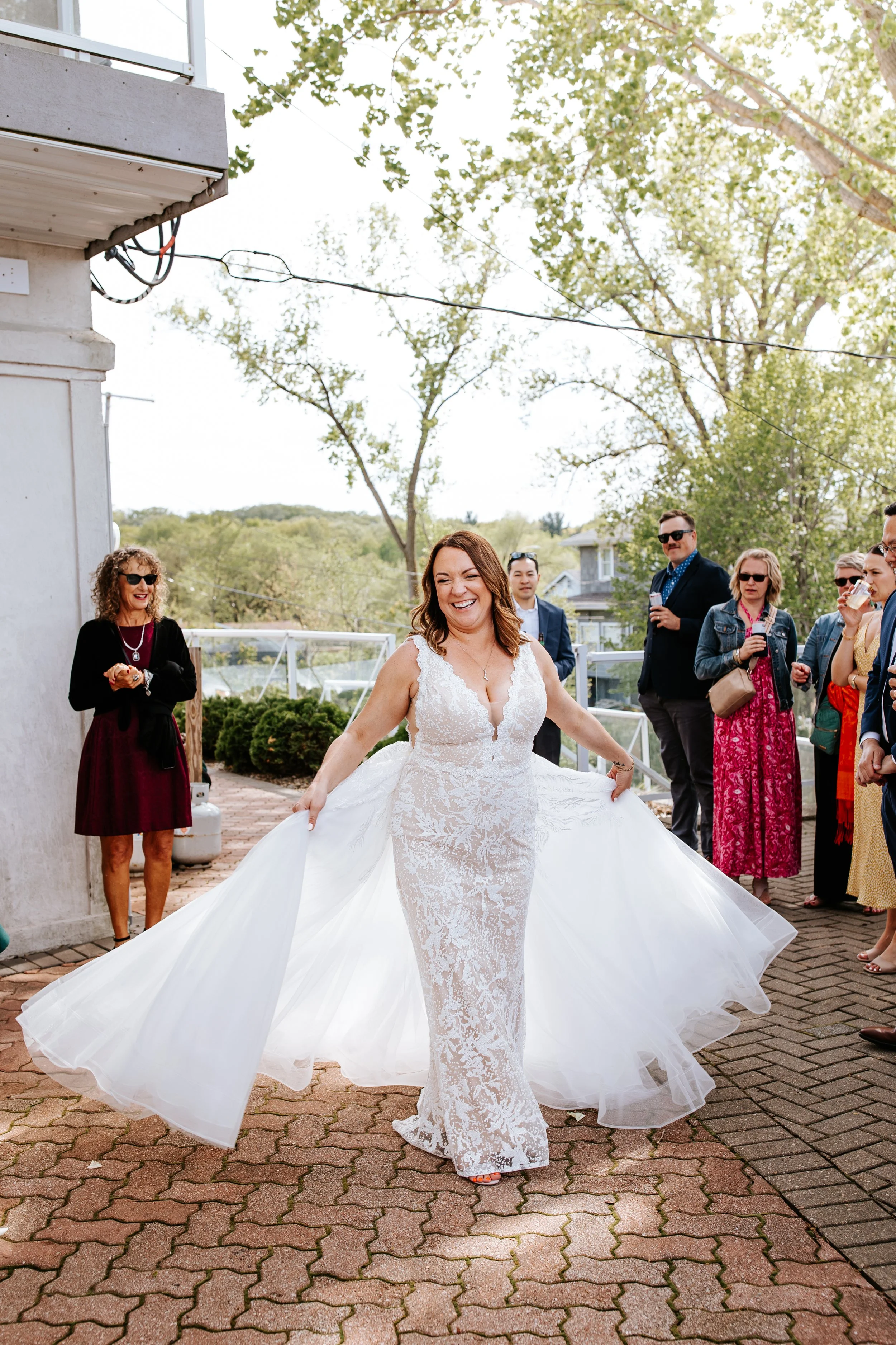 A bride in a white lace wedding dress smiling and holding the sides of her train outside surrounded by friends and family on a sunny day.