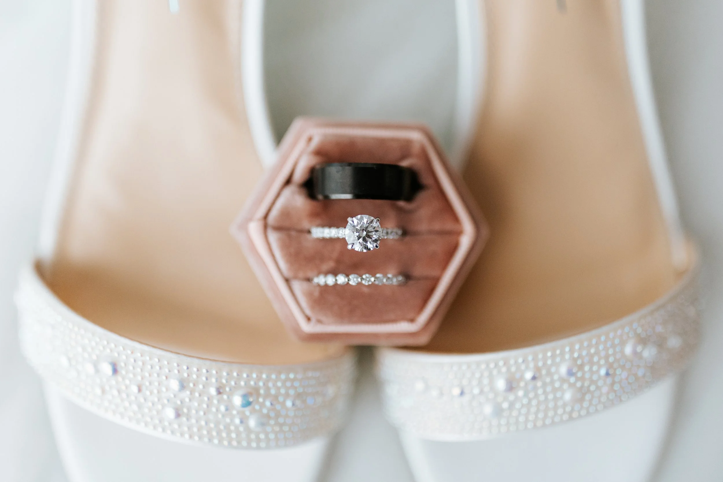 Close-up of a hexagonal pink ring box with three rings inside: an engagement ring with a large central diamond, a black band, and a diamond-studded band, on a white garment with pearl and crystal embellishments.