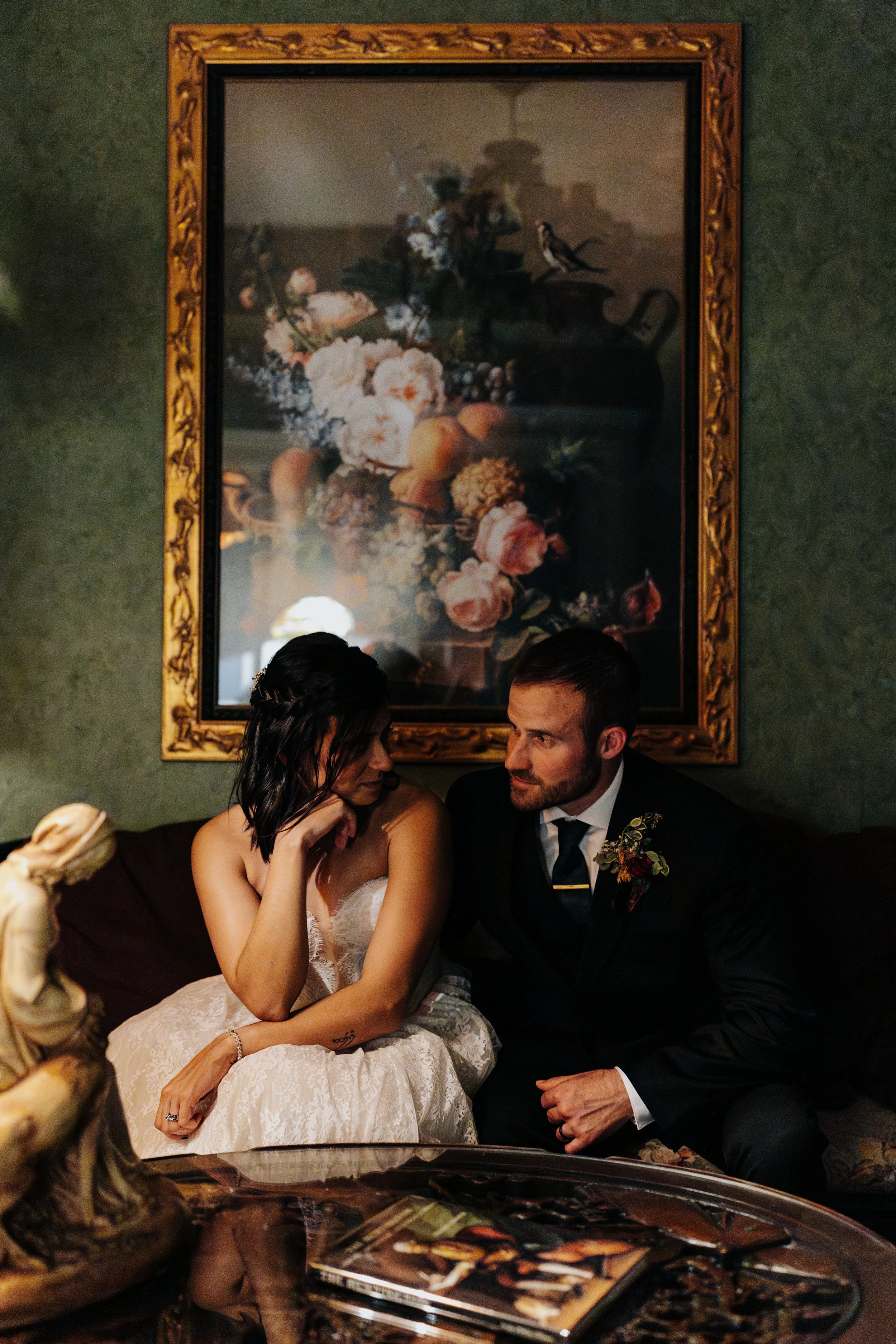 A bride and groom sitting on a vintage sofa in a dimly lit room, engaged in an intimate conversation, with a large framed floral painting behind them.