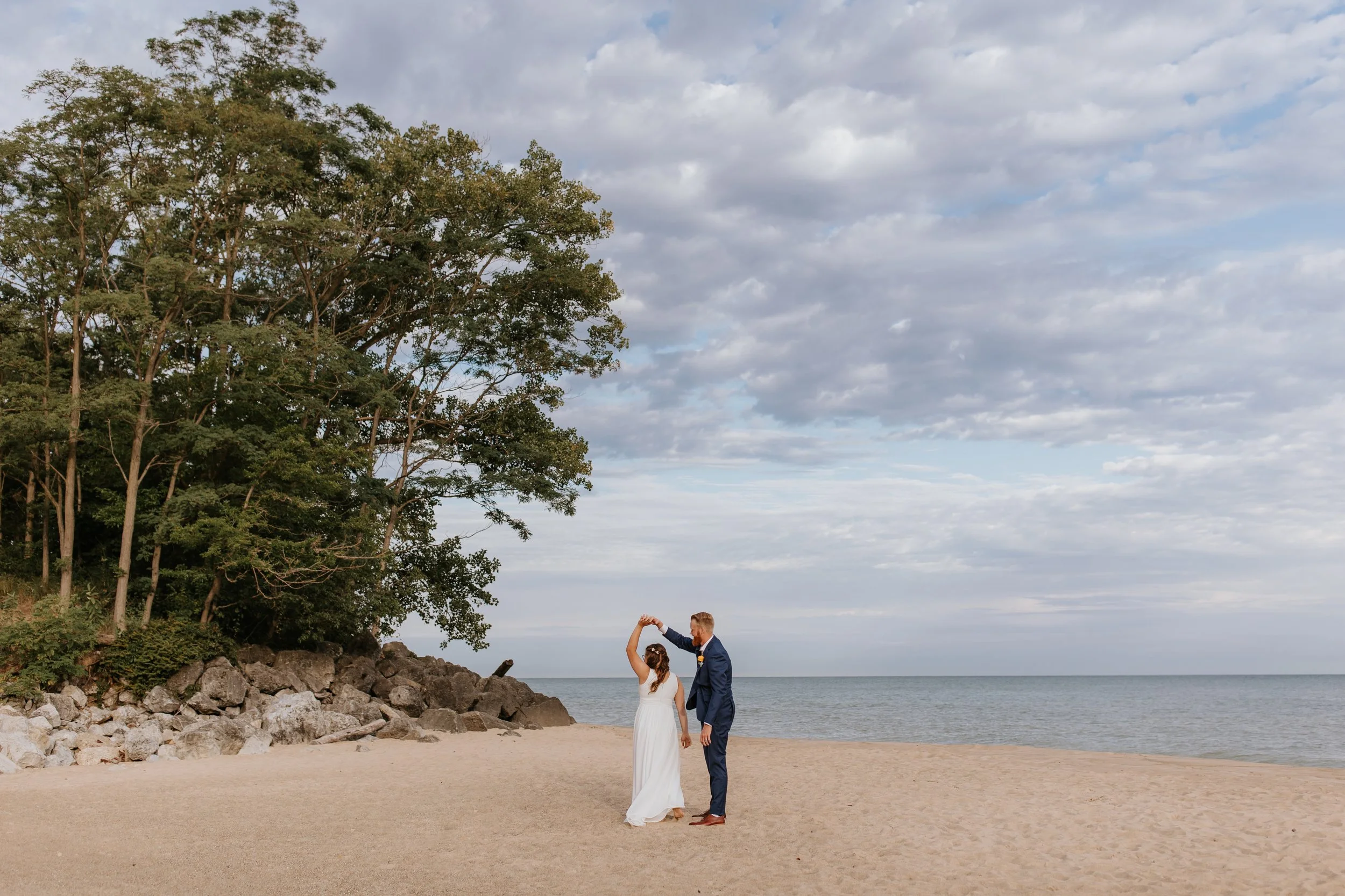 A bride and groom dancing on a beach with a rocky shoreline, trees, and a cloudy sky in the background.