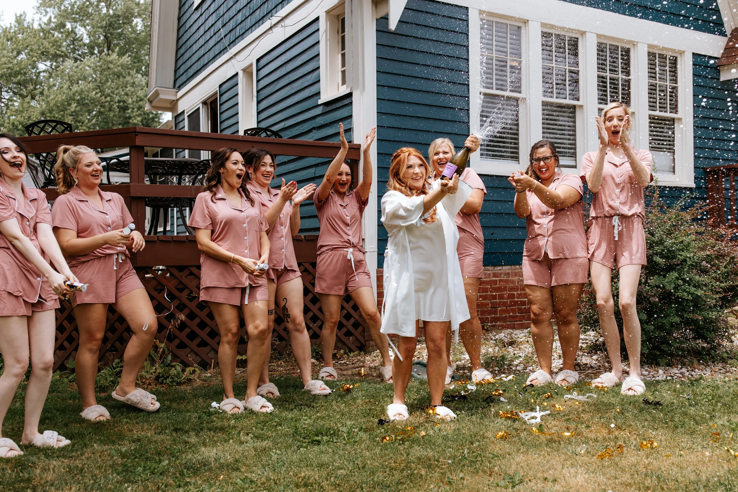 A group of women in matching pink pajamas celebrating outdoors with a woman in white spraying champagne as others cheer and laugh.