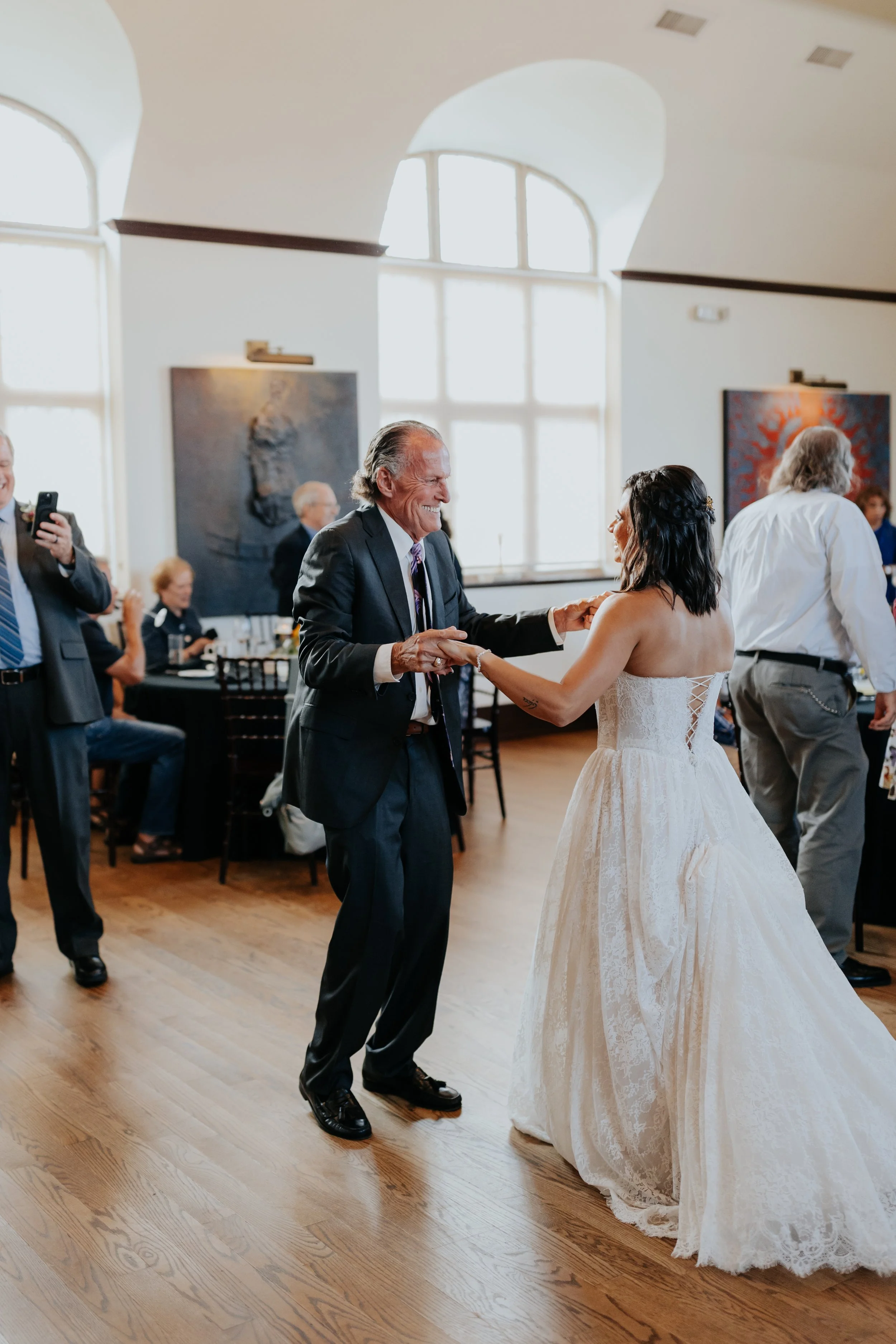 A bride in a white wedding gown dances with an older man in a suit at a wedding reception. Other guests are seated or standing in the background, some taking photos, in a bright room with large windows and wooden floors.