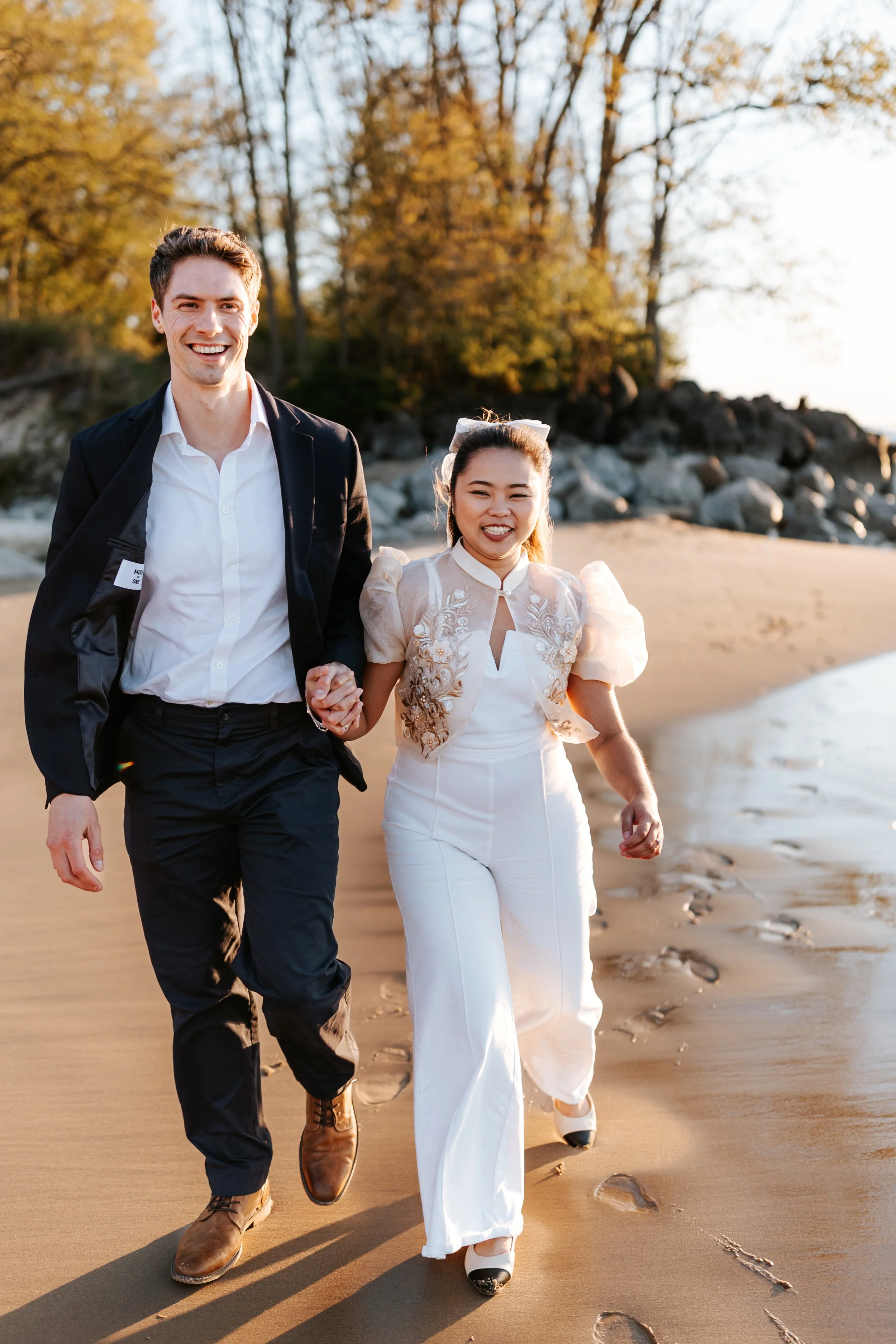A happy couple walking hand-in-hand on the beach, late afternoon or early evening, with fallen leaves and autumn trees in the background.