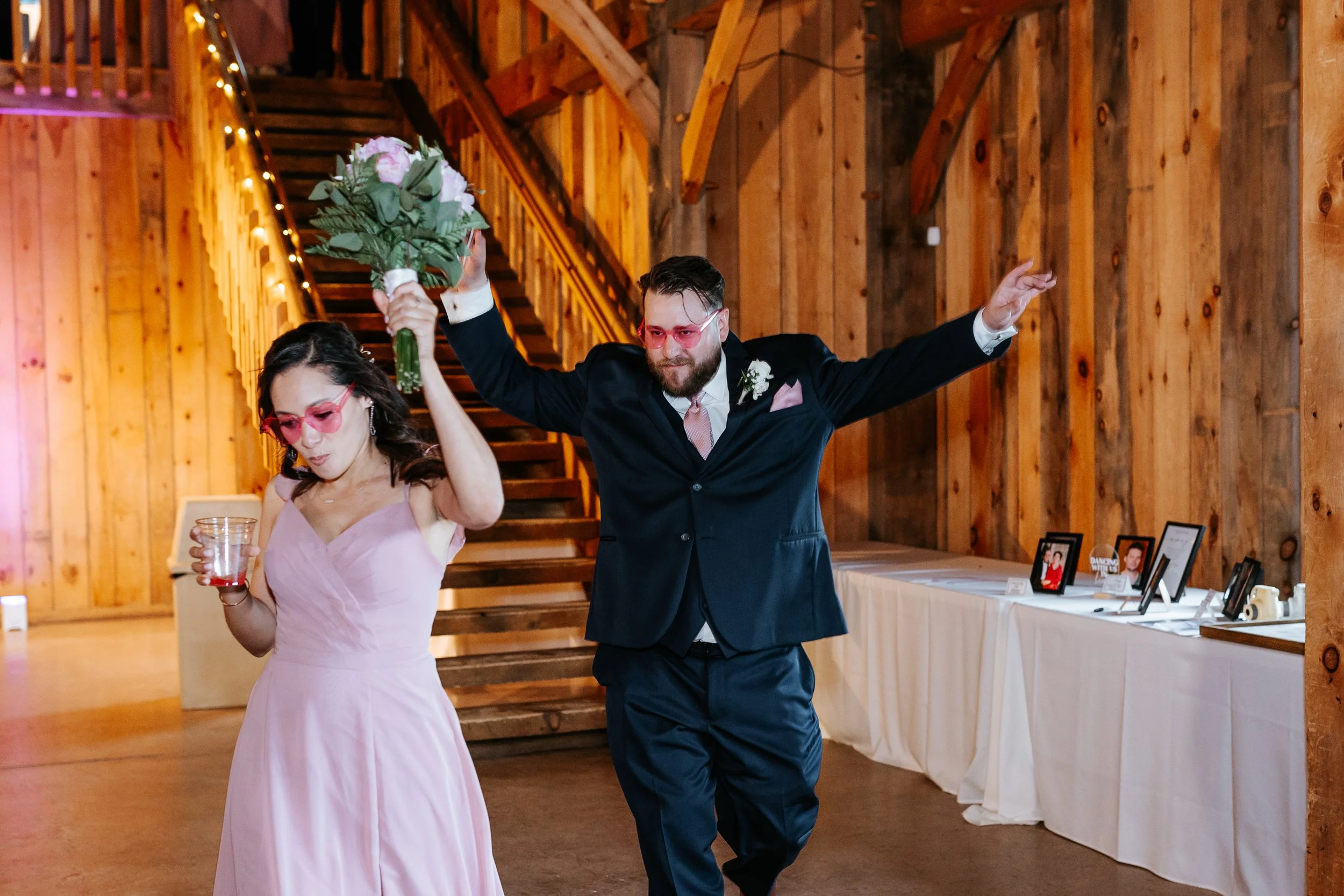 A man in a black suit with pink-tinted sunglasses holding a bouquet of pink and white flowers, dancing with a woman in a pink dress wearing pink-tinted sunglasses at a wedding reception with wooden walls and a staircase in the background.
