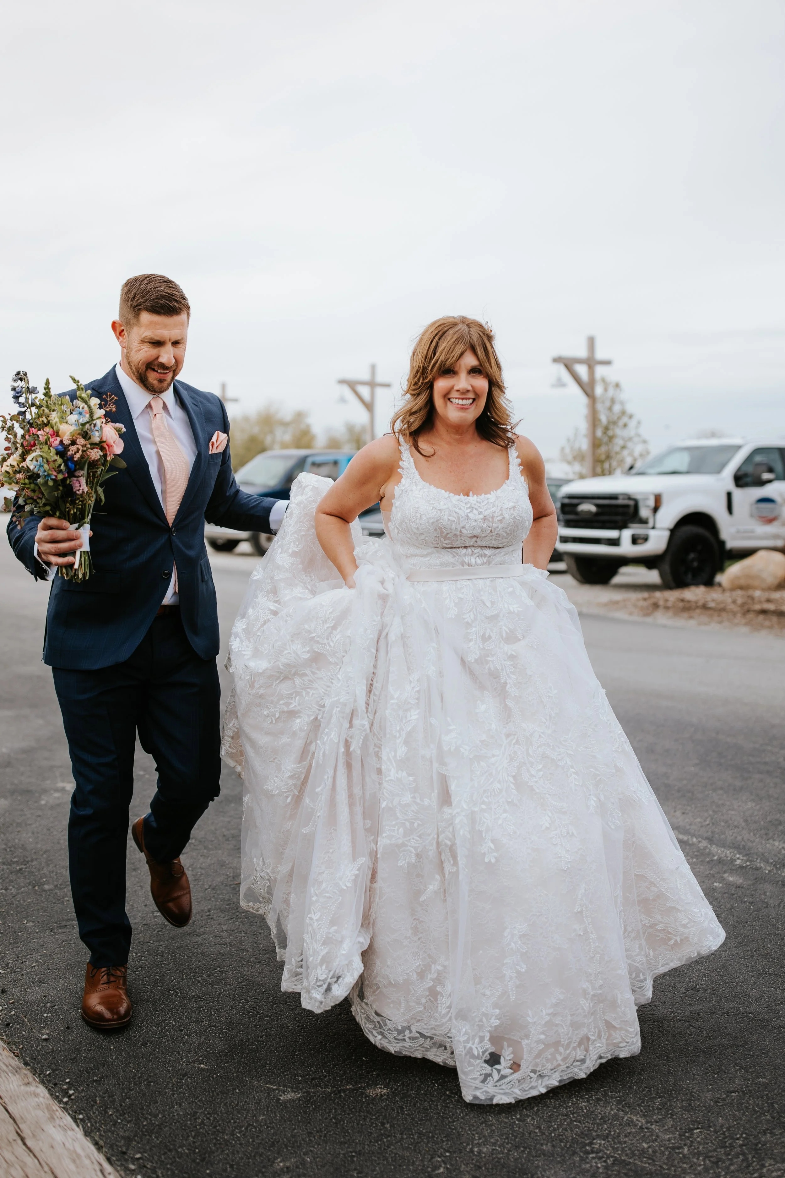 A bride in a white lace wedding dress smiling and holding up her gown, accompanied by a groom in a navy suit holding a bouquet, walking outdoors on a paved road with vehicles in the background.