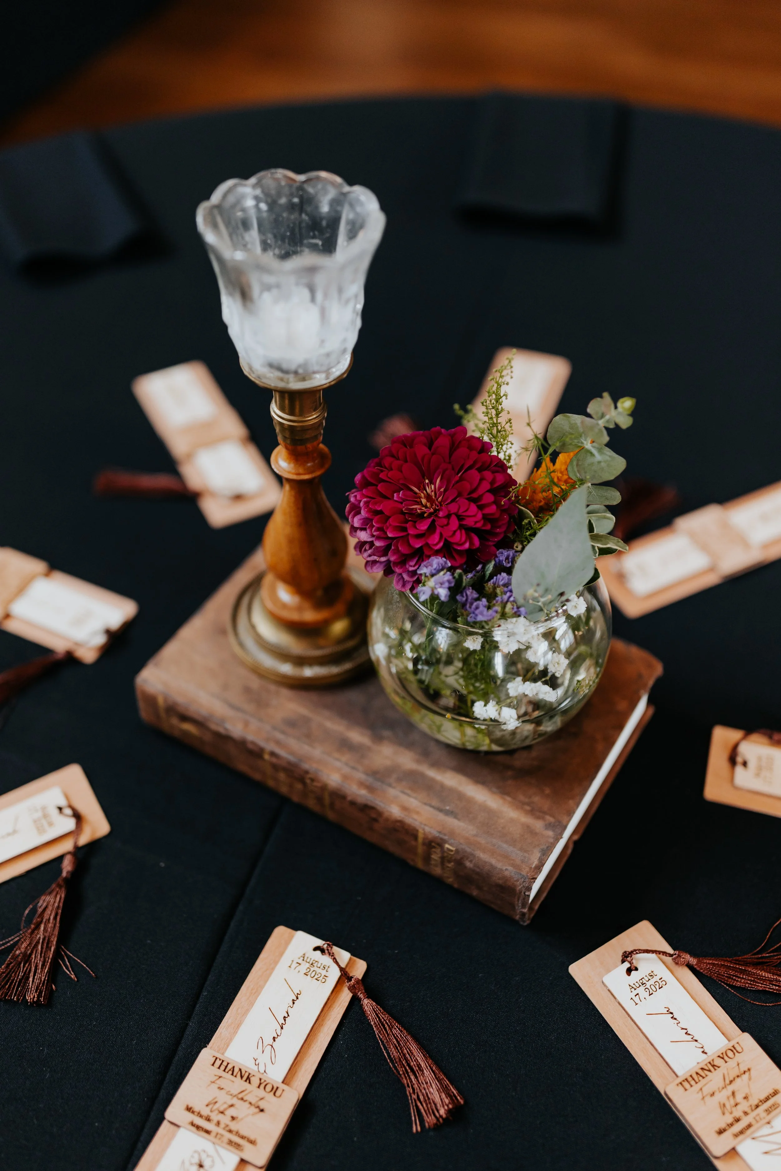 A black table with a centerpiece consisting of a vintage glass candle holder, a wooden candlestick, and a small glass bowl with colorful flowers, placed on a couple of old books. Surrounding the centerpiece are tan place cards with handwritten names,
