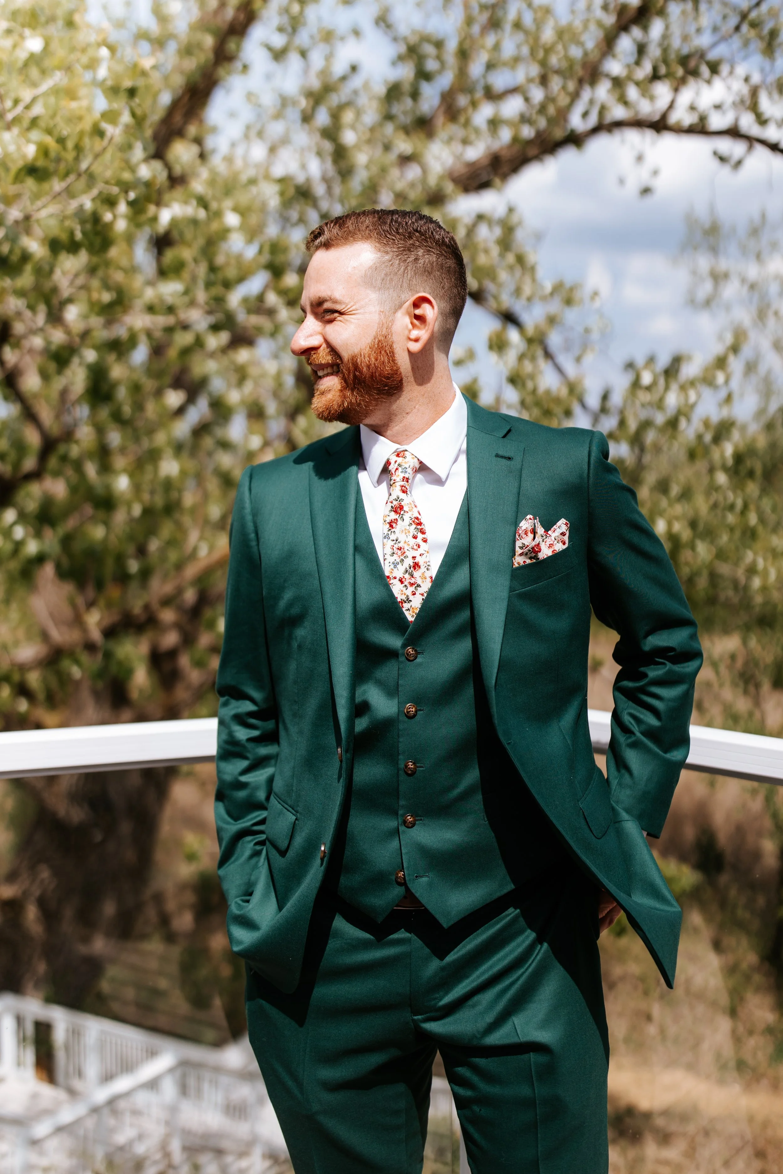 A man in a green suit smiling and looking to his left outdoors with trees and blue sky in the background.
