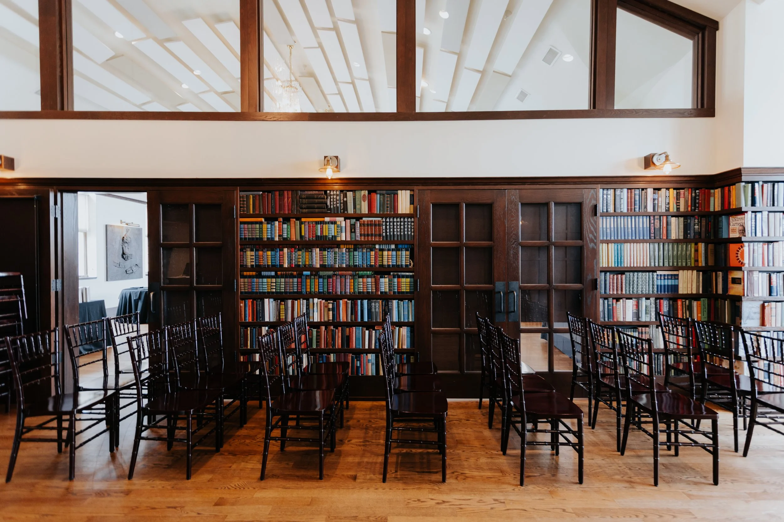 Interior of a library with wooden bookshelves filled with books, black chairs arranged in rows, wooden flooring, and a upper-level balcony with glass windows.