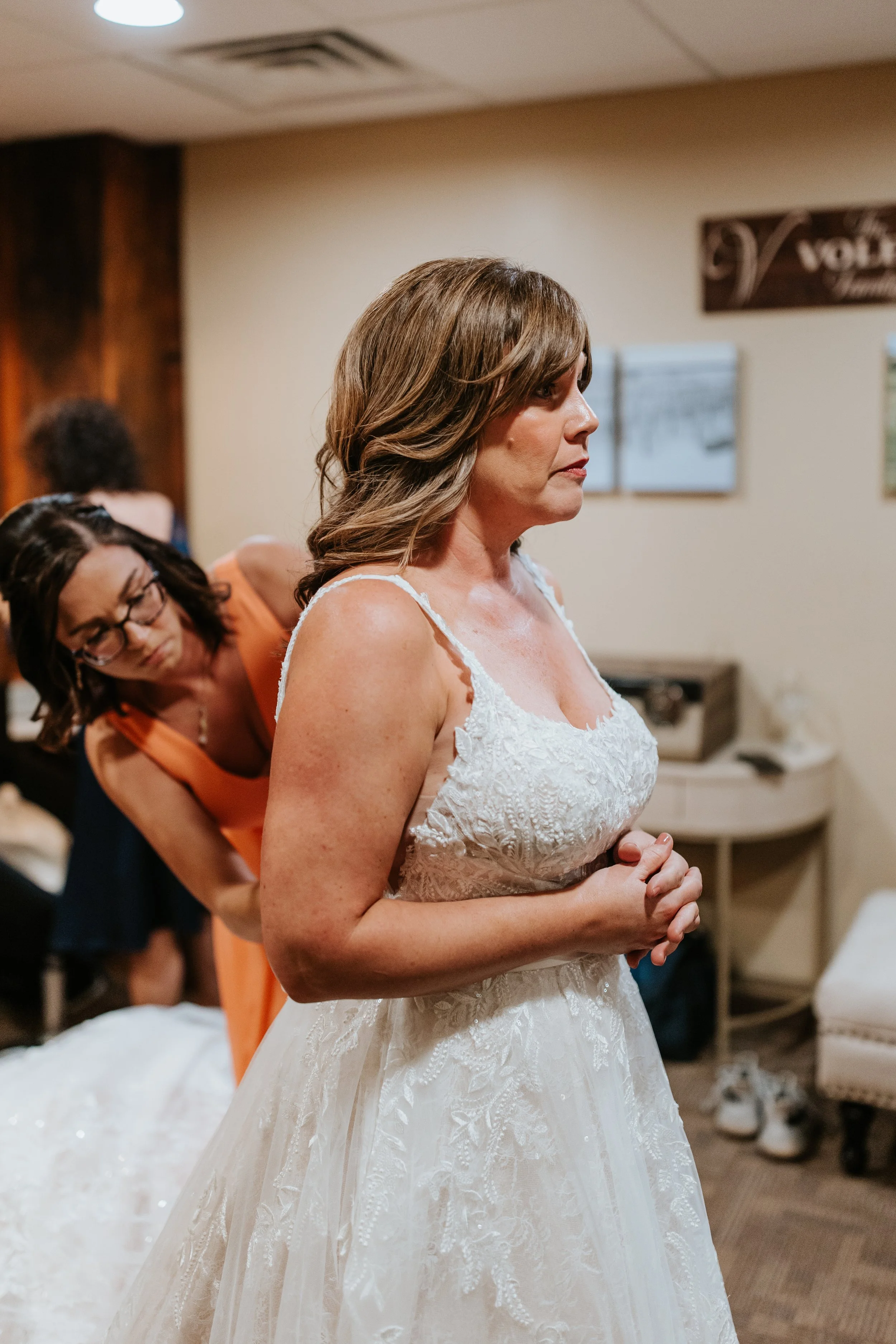 A woman in a white lace wedding dress standing with her hands clasped together, with another woman behind her helping her with her dress in a room with beige walls and wooden accents.