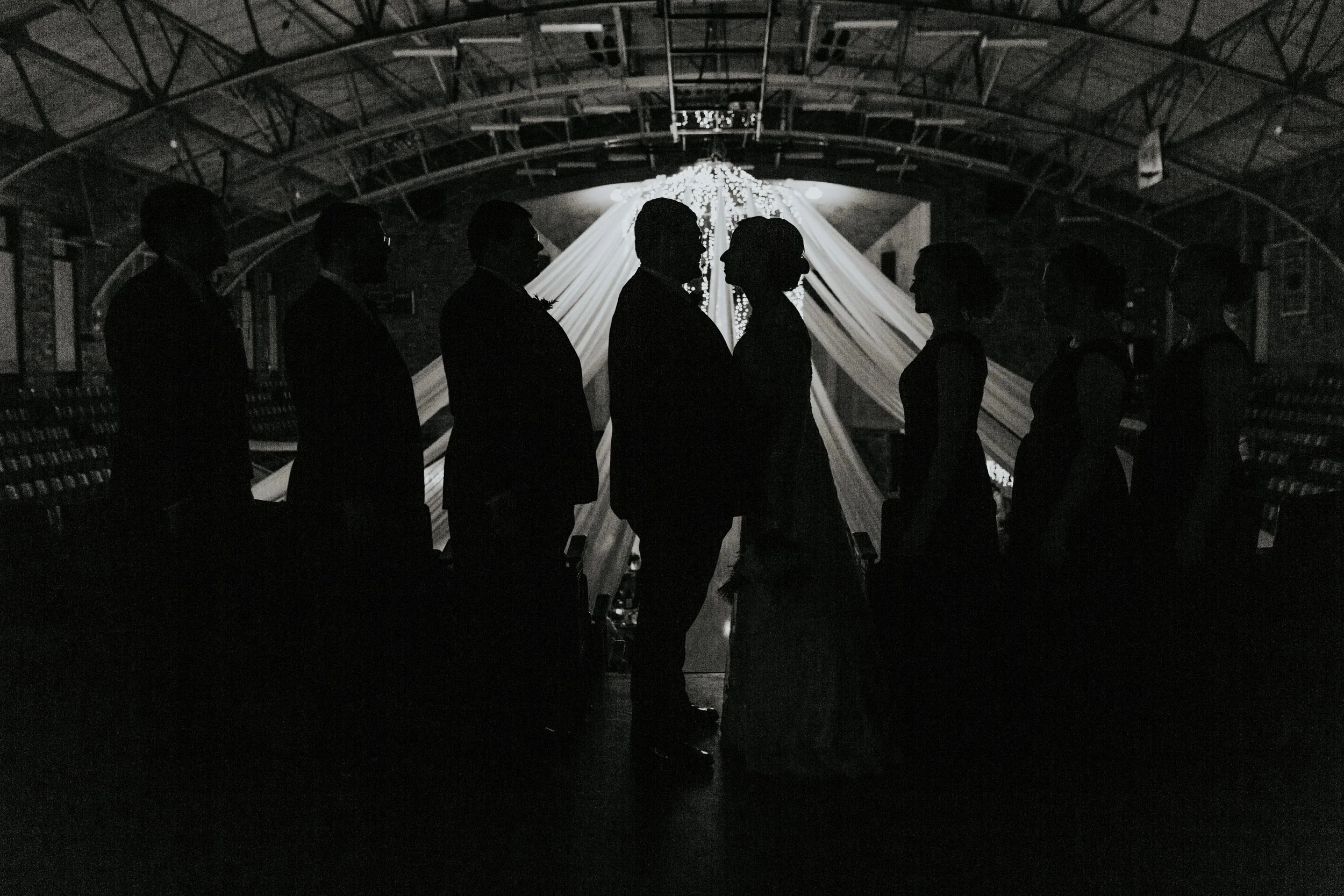 Silhouettes of a couple with their wedding party in an indoor venue with draped fabric and a chandelier in the background, capturing a romantic moment.
