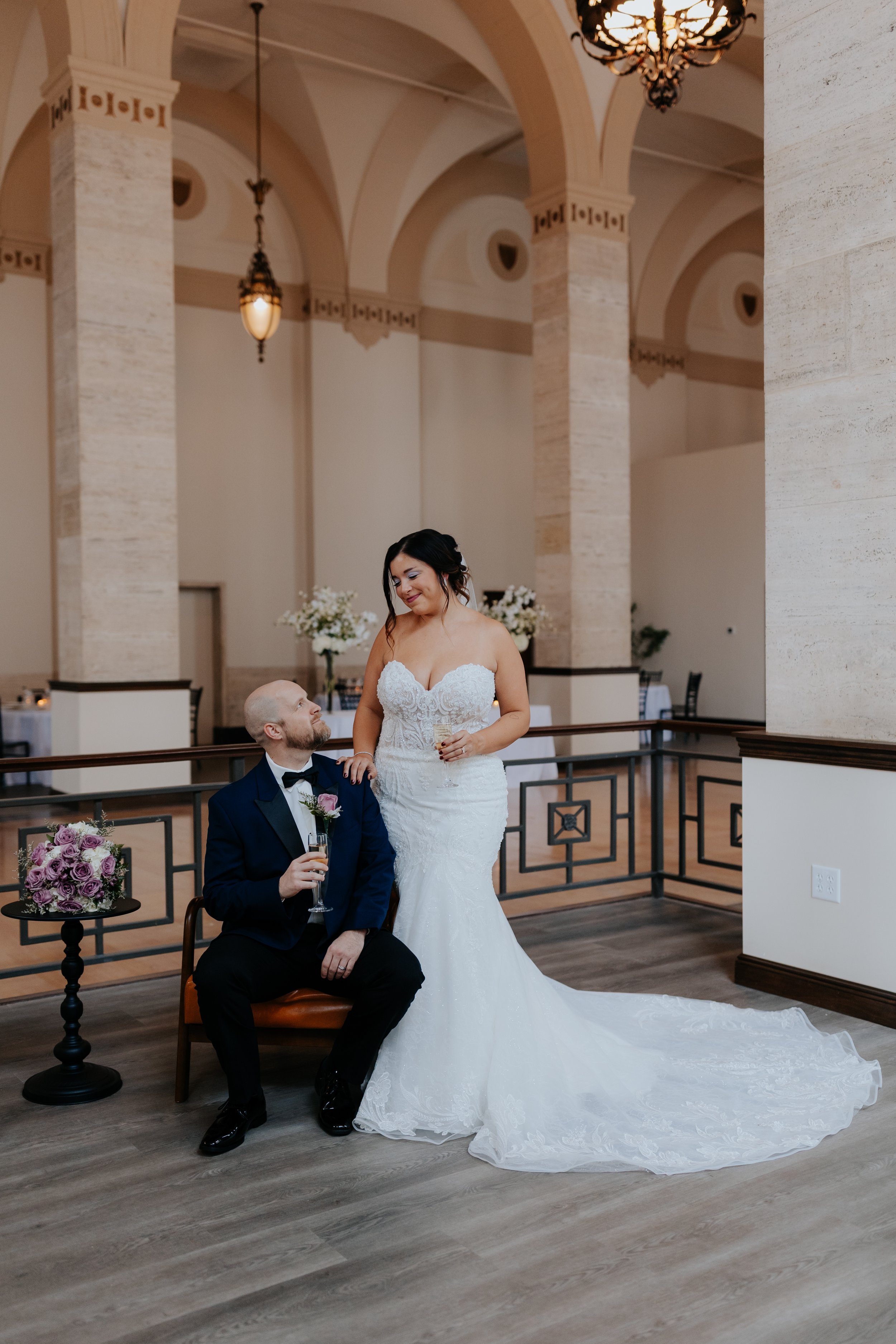 Bride and groom at their wedding reception, with the bride in a white lace wedding gown and the groom in a navy tuxedo sitting on a chair, holding champagne glasses in a decorated venue.