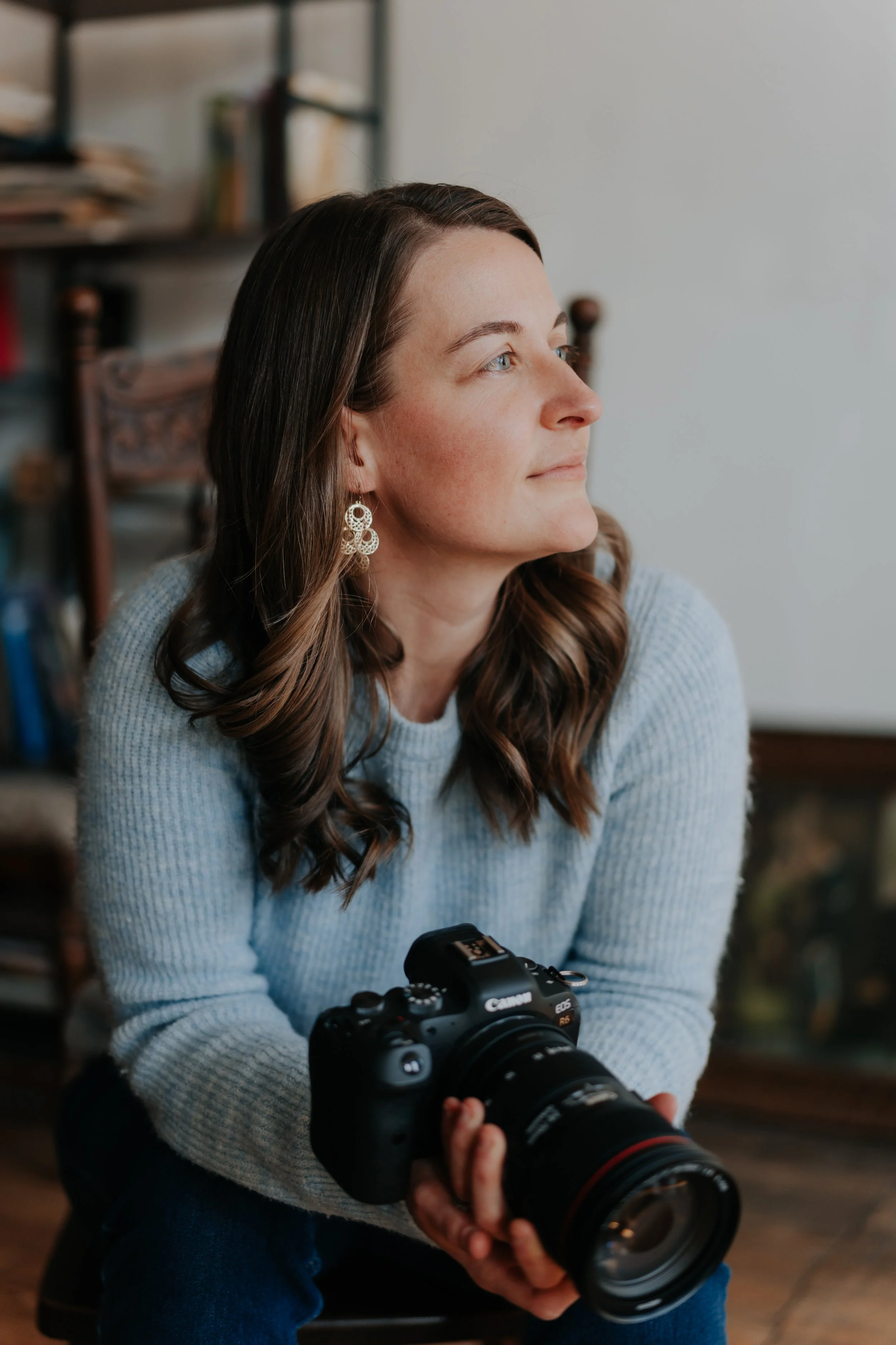A woman with long brown hair and earrings holding a Canon camera, sitting indoors.
