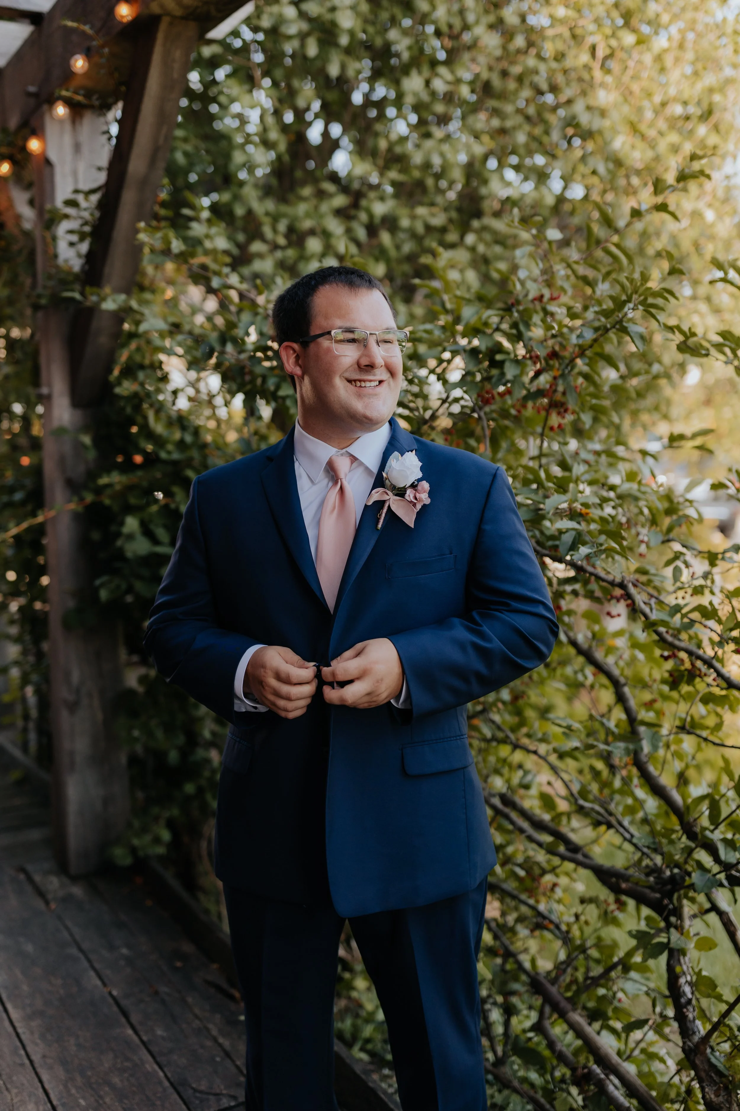 A man in a navy suit with a pink tie and a boutonniere, smiling and looking to the side, standing outdoors on a wooden pathway with greenery in the background.