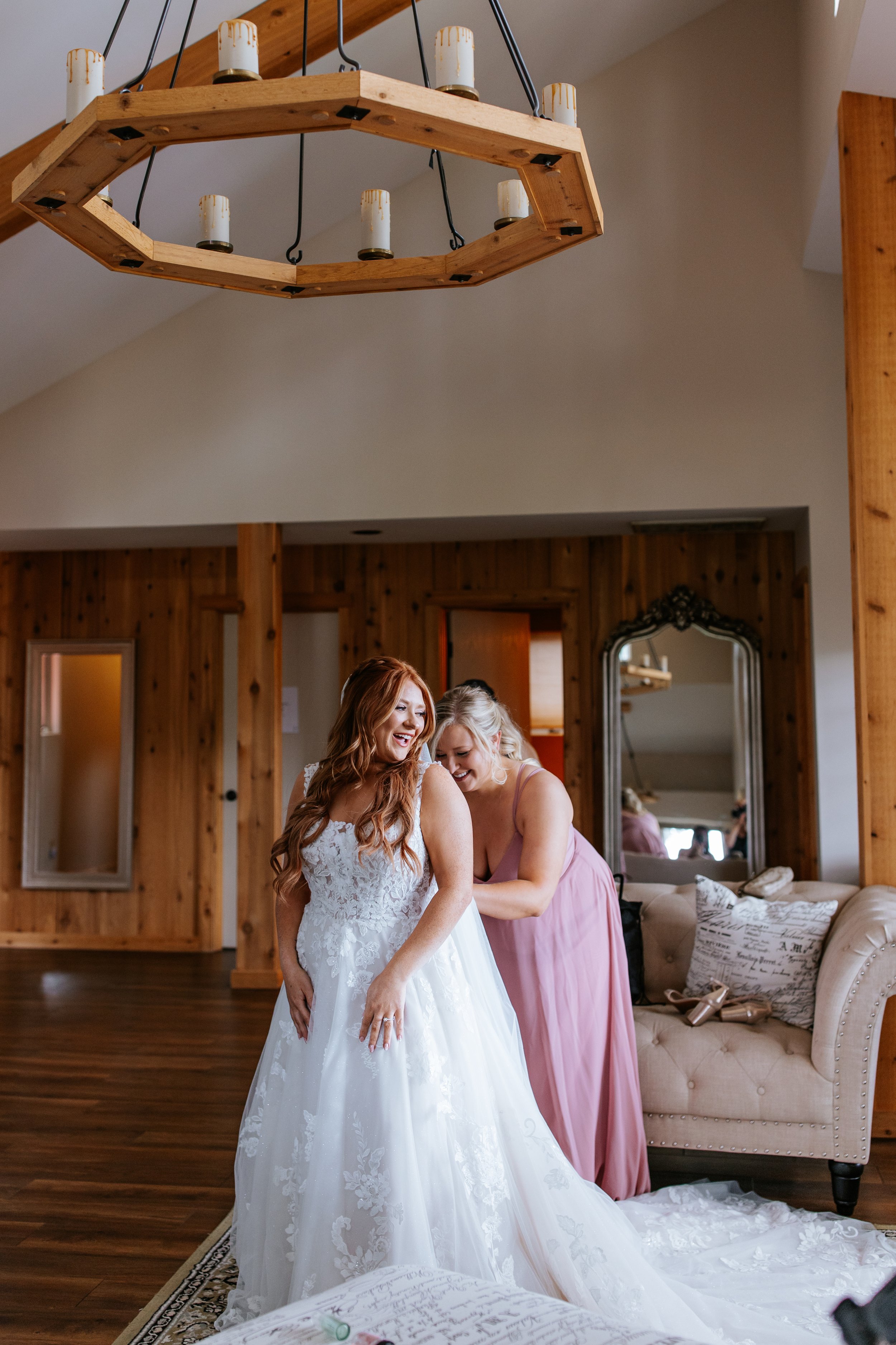 A bride in a white lace wedding gown smiling as a woman in a pink dress helps her get ready, inside a cozy room with wooden walls and furniture.