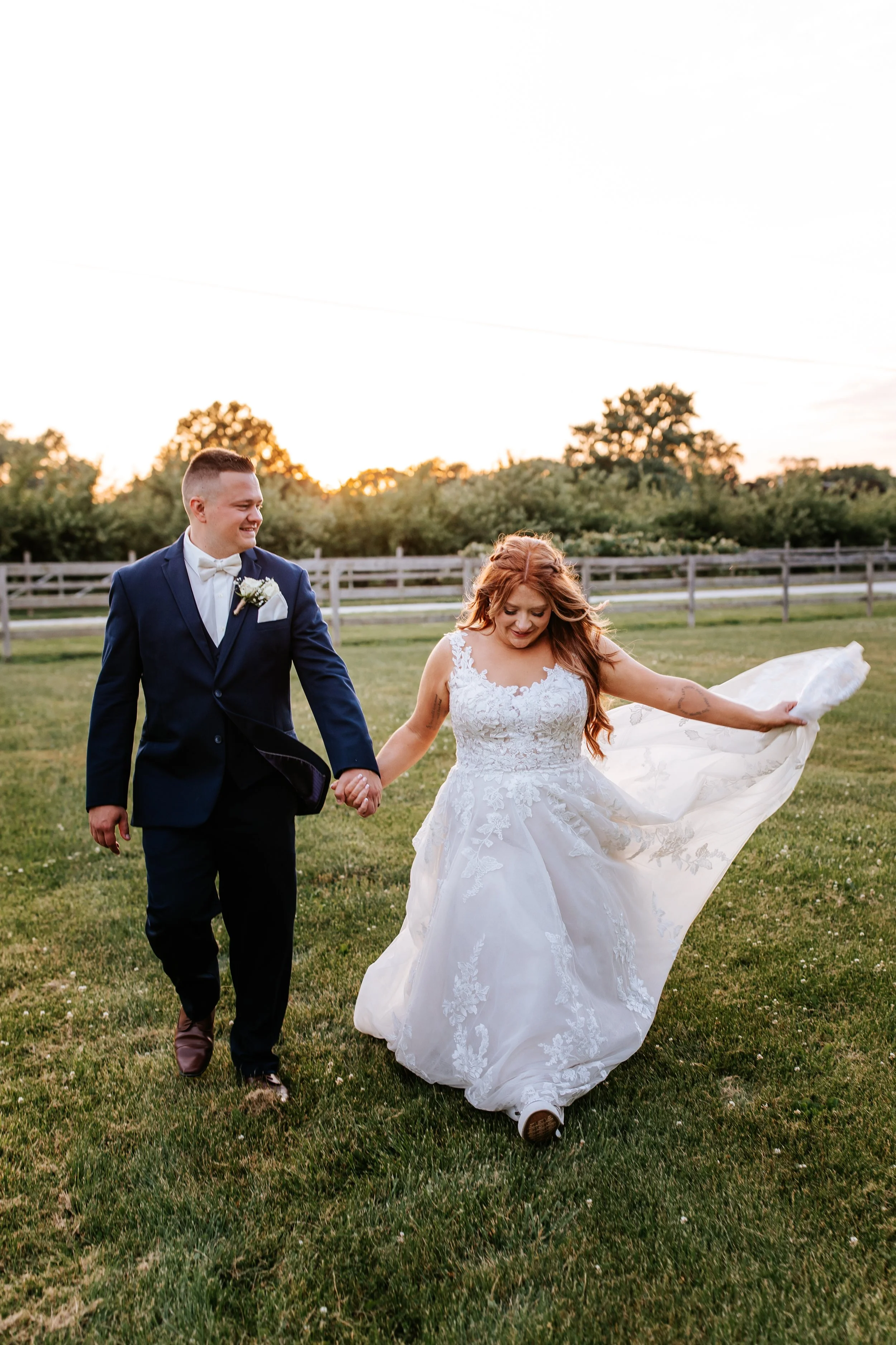 A newlywed couple holding hands and walking on a grassy field during sunset, with trees and a white fence in the background.