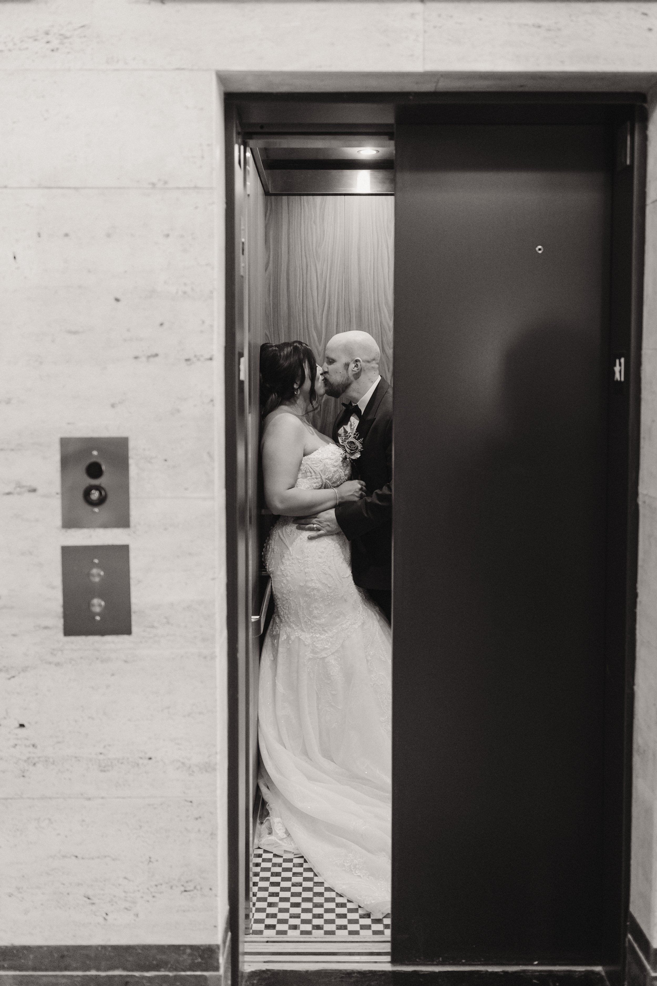 Black and white photo of a couple dressed in wedding attire sharing a kiss inside an elevator.