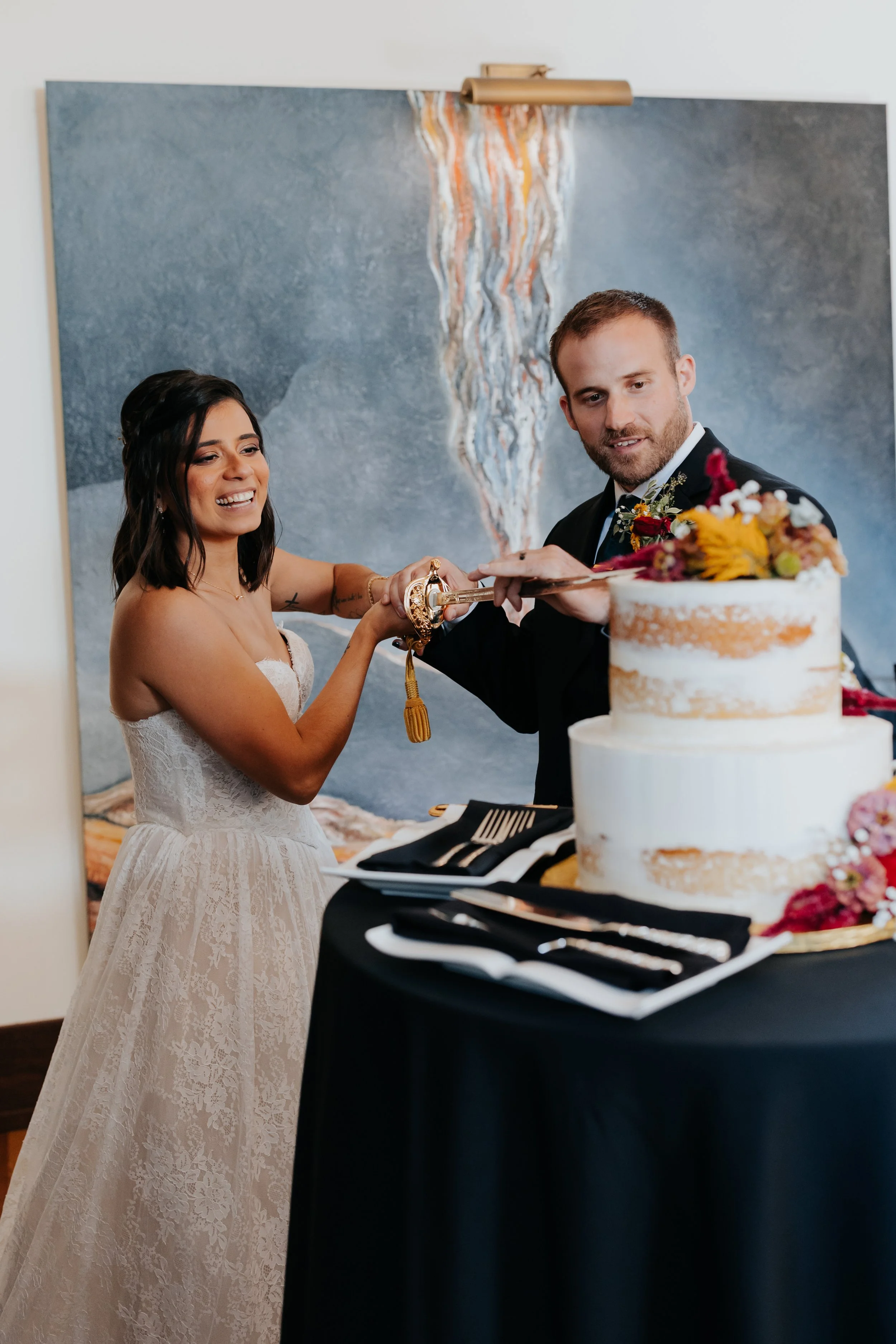A bride and groom cutting a wedding cake together, smiling and holding a sword.
