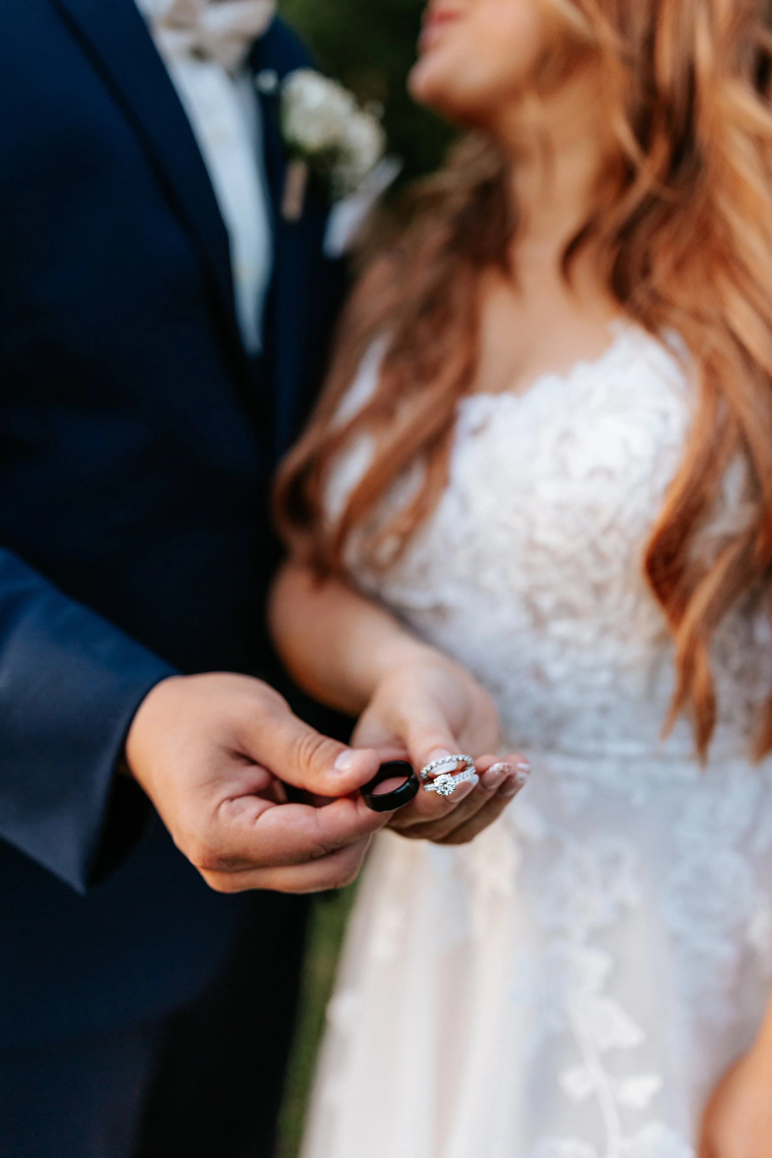 A couple at their wedding, with the groom in a black suit holding a wedding ring and the bride in a white lace dress.