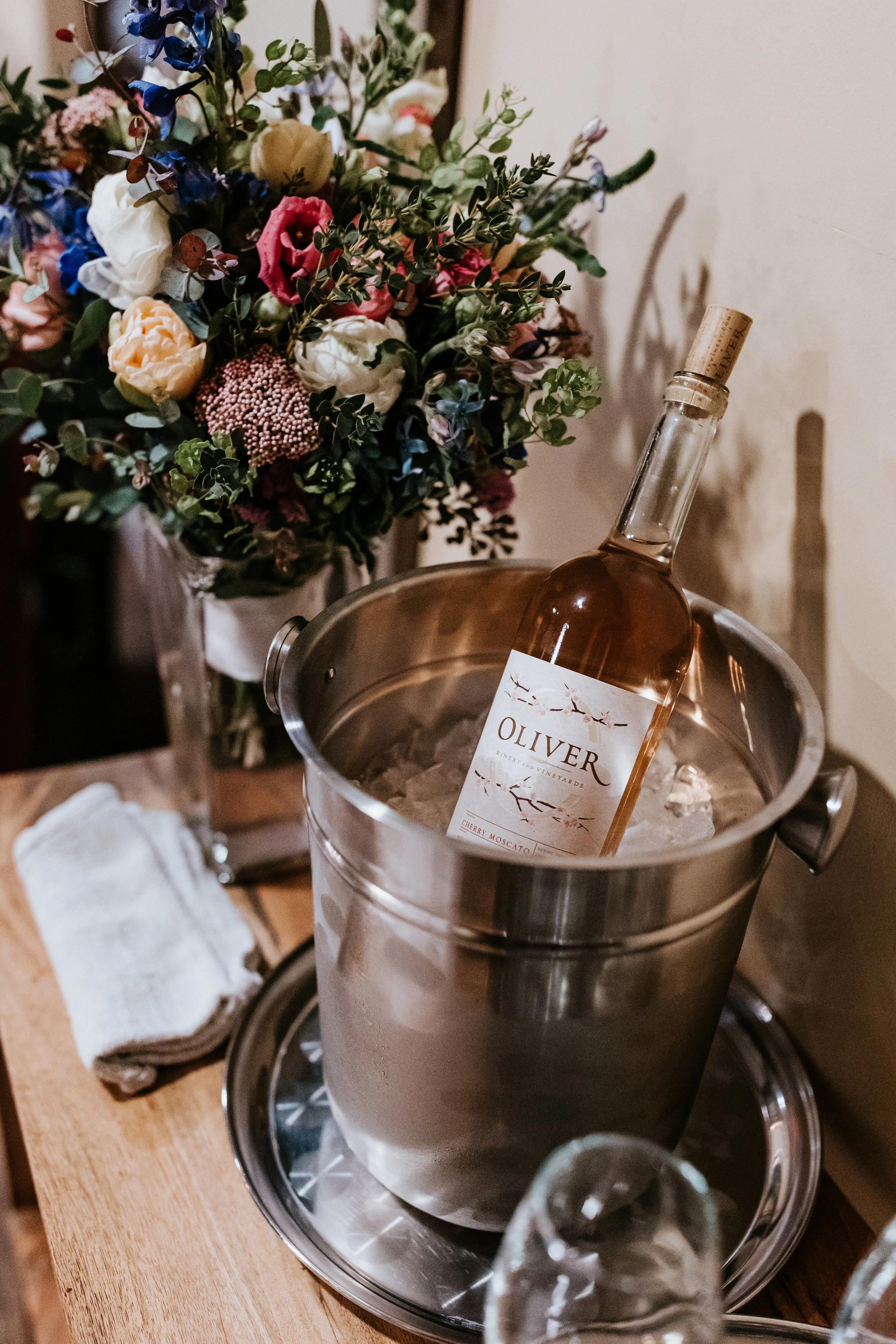 A bottle of Oliver cherry Moscato wine in an ice bucket, with a bouquet of colorful flowers in the background.