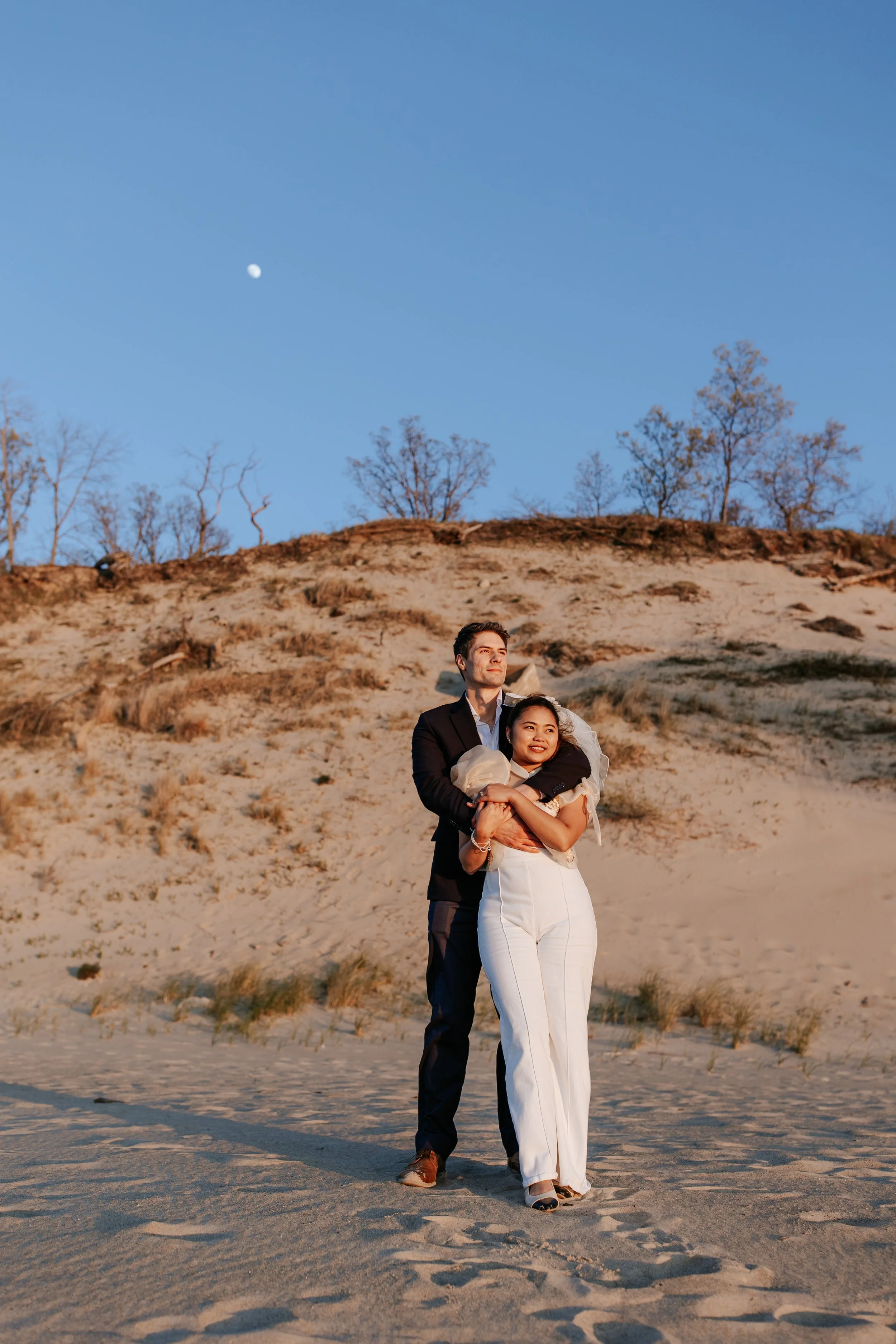 A couple dressed in wedding attire standing on a sandy beach at sunset with a hill and sparse trees in the background, and the moon visible in the sky.
