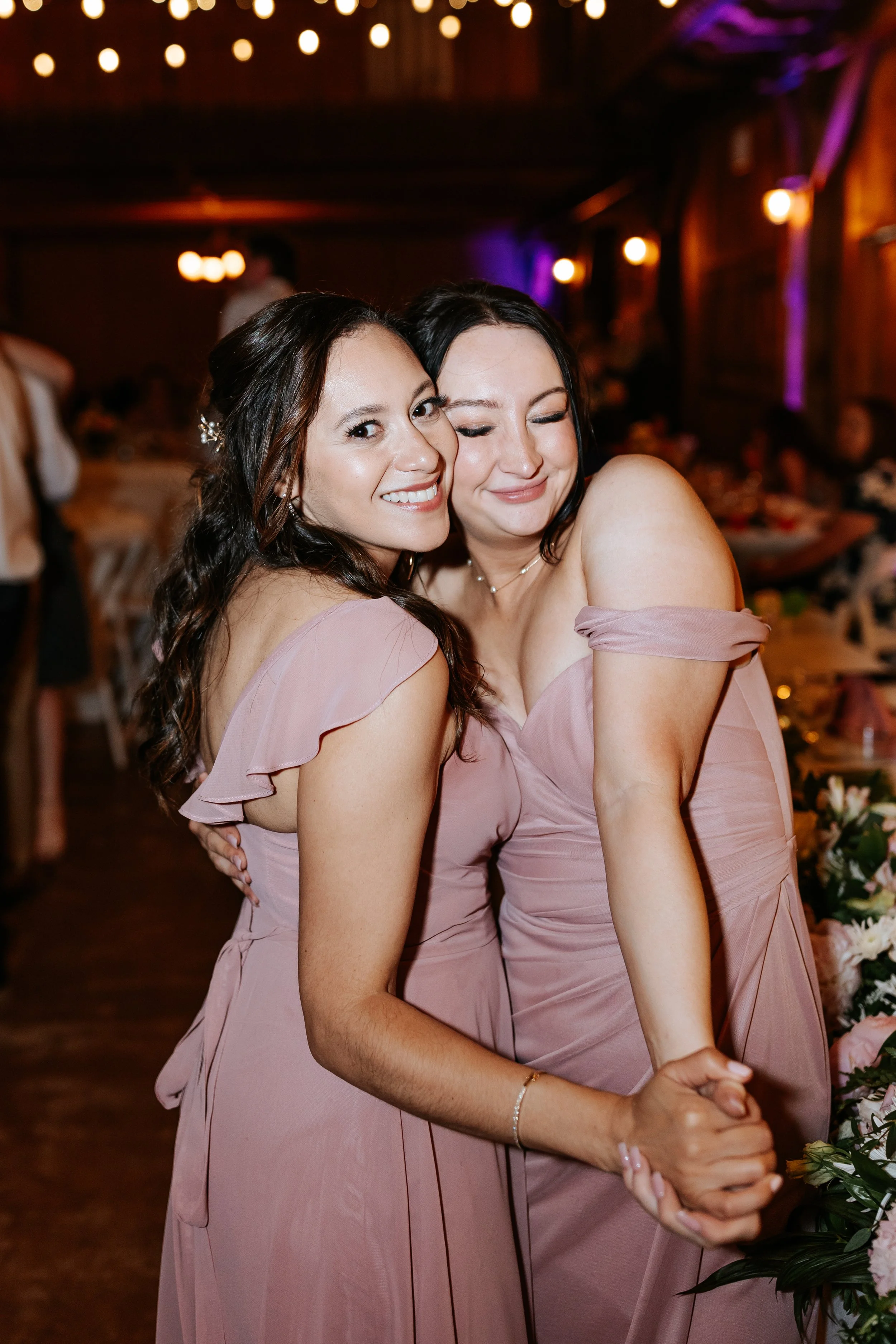 Two women in pink bridesmaid dresses hugging and smiling at a wedding reception.