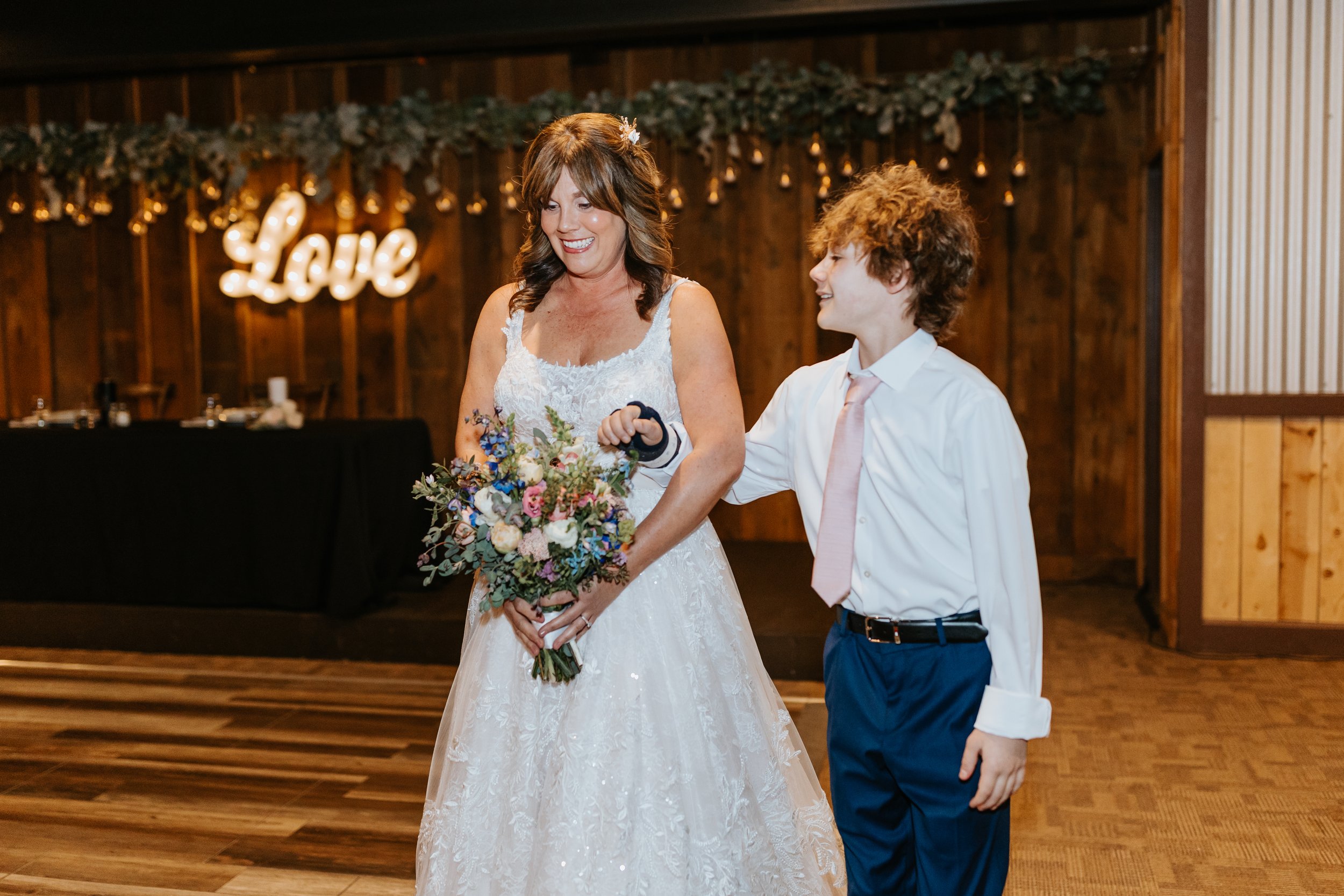 A woman in a white wedding dress holding a bouquet of flowers smiling at a young boy in a white shirt and pink tie at a wedding reception with a wooden backdrop and illuminated 'Love' sign.